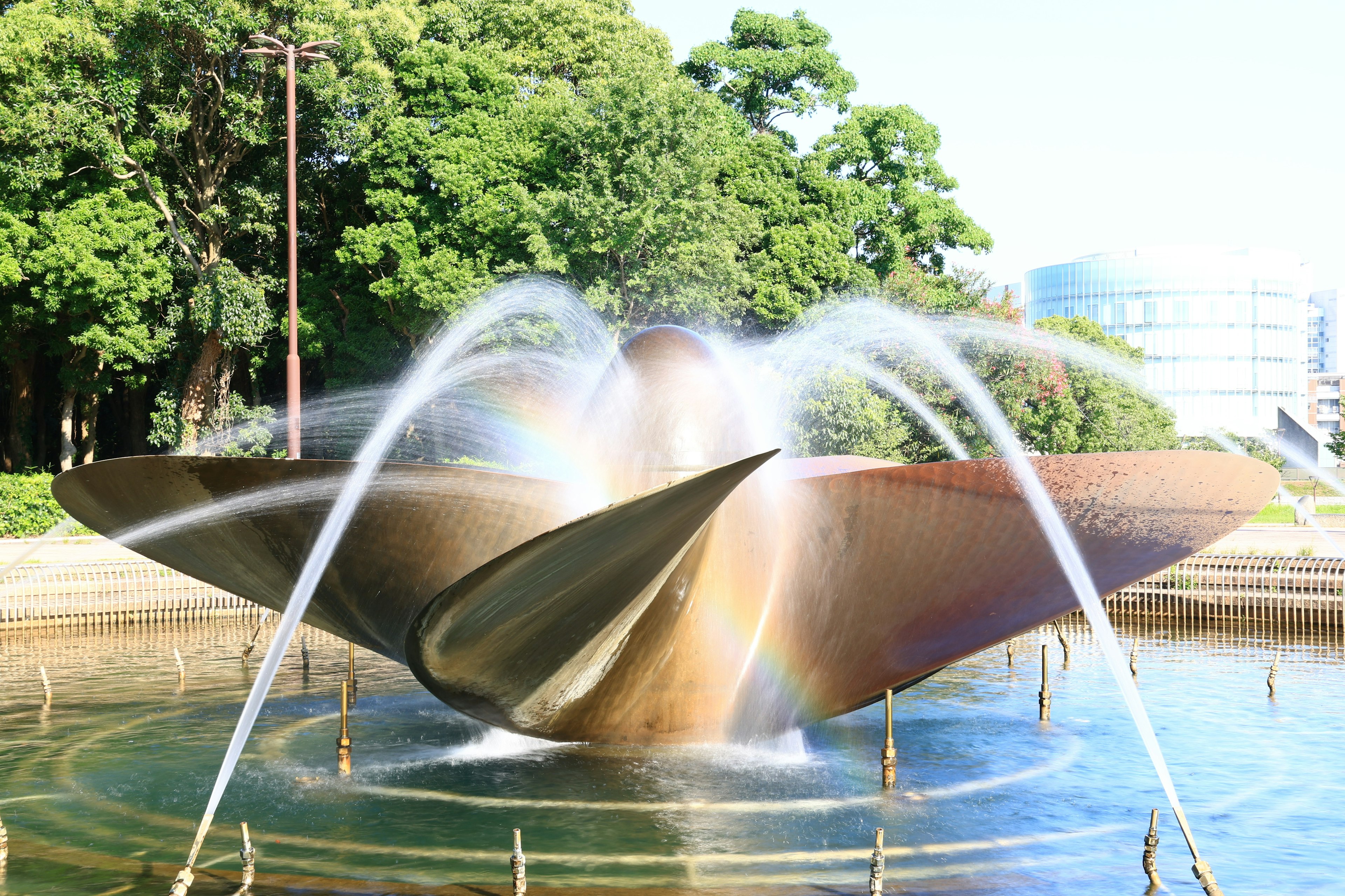 A beautiful metallic fountain at the center of a park with flowing water and surrounding green trees