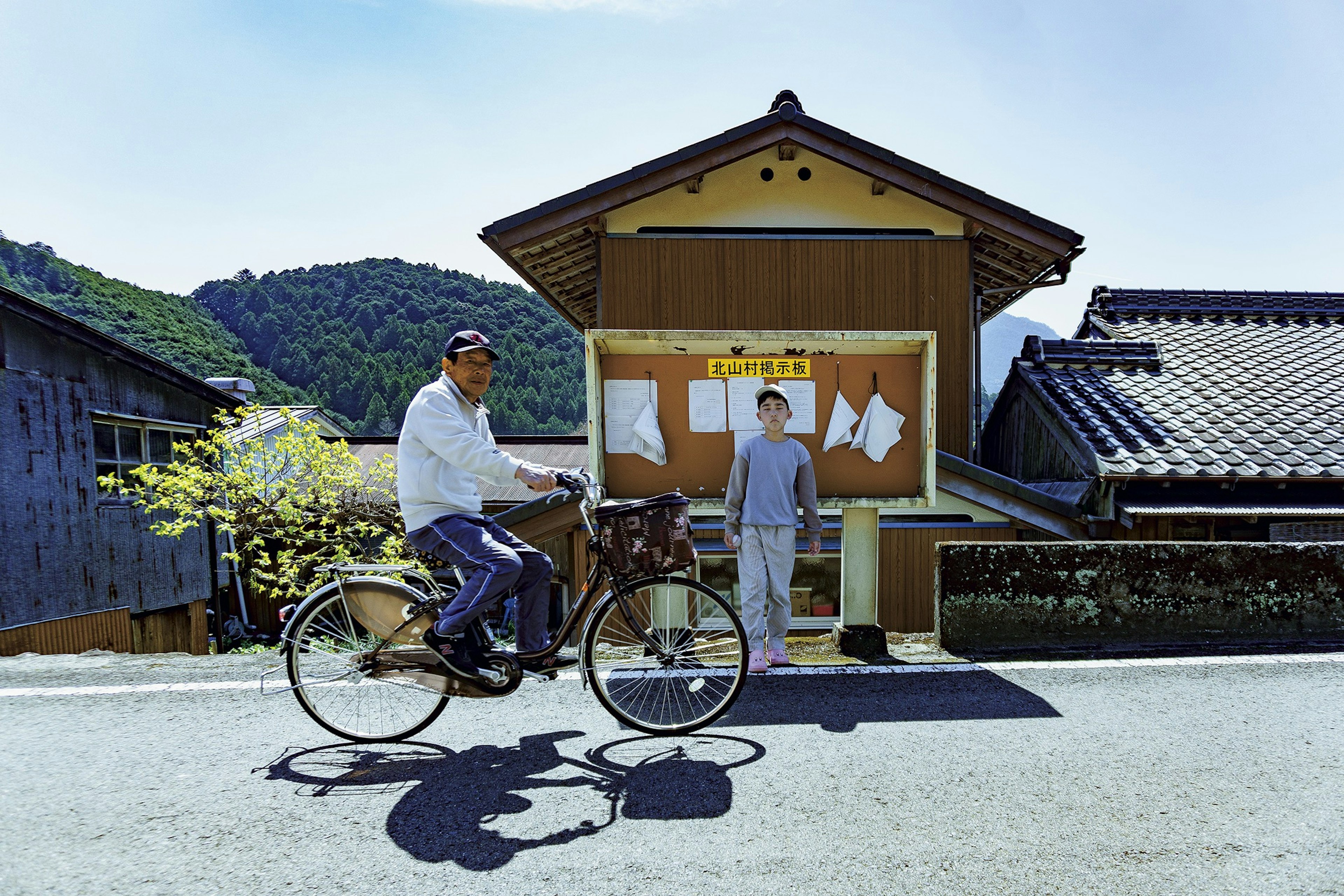 Un hombre mayor montando una bicicleta con una mujer de pie cerca de una casa japonesa tradicional