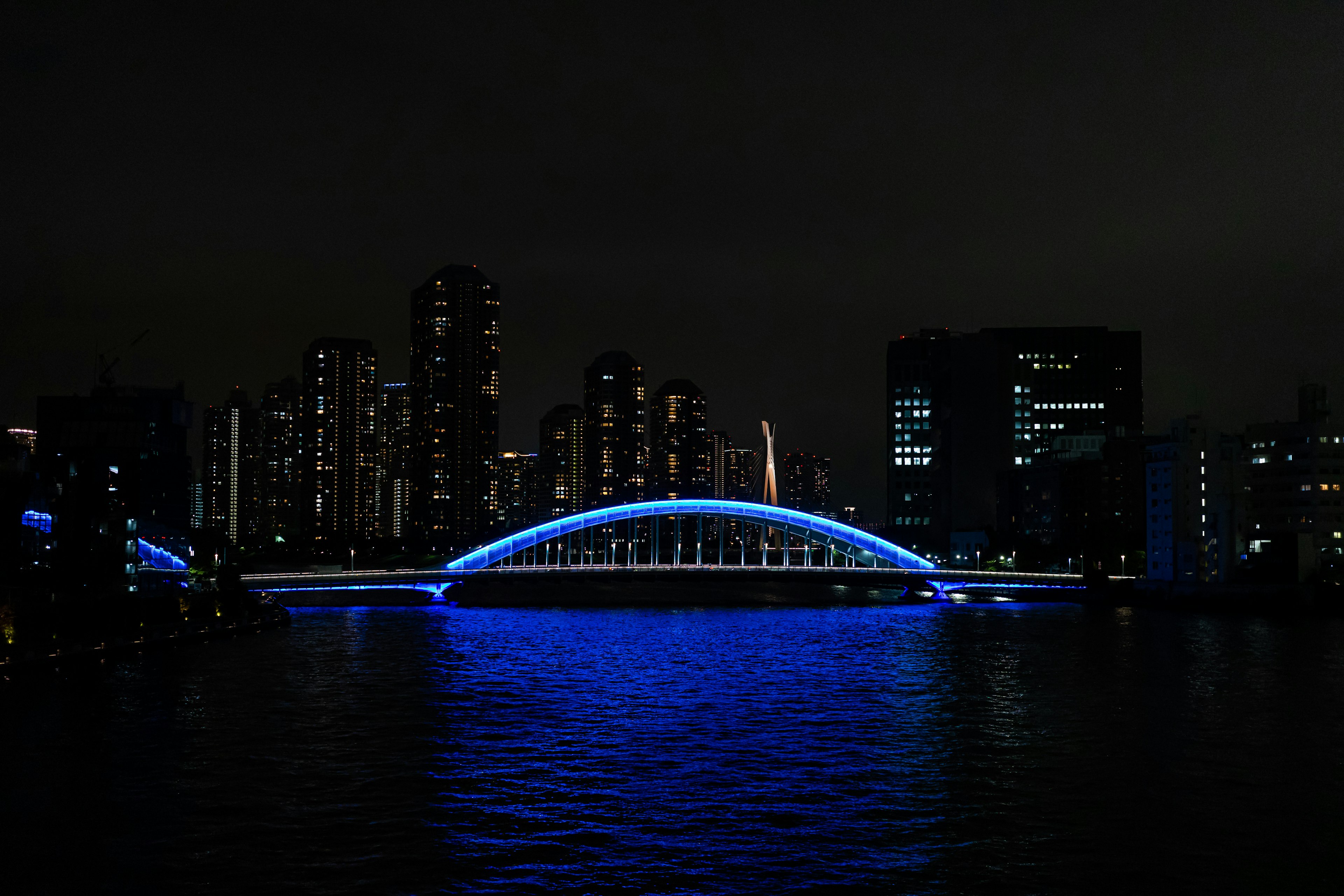 Jembatan lengkung yang diterangi lampu biru di atas sungai dengan cakrawala kota di malam hari