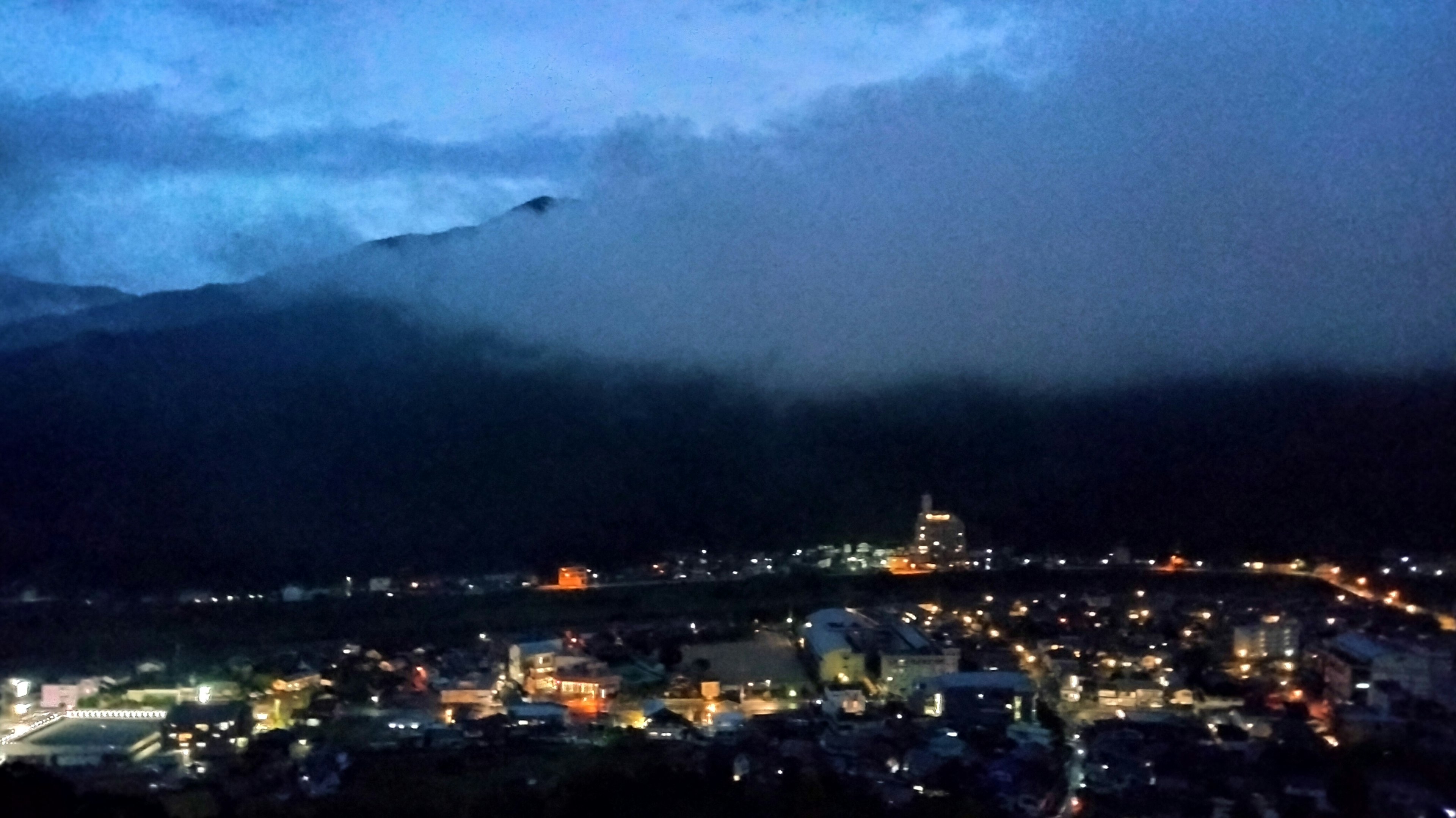 Vista notturna di uno skyline cittadino con montagne in silhouette avvolte da nuvole