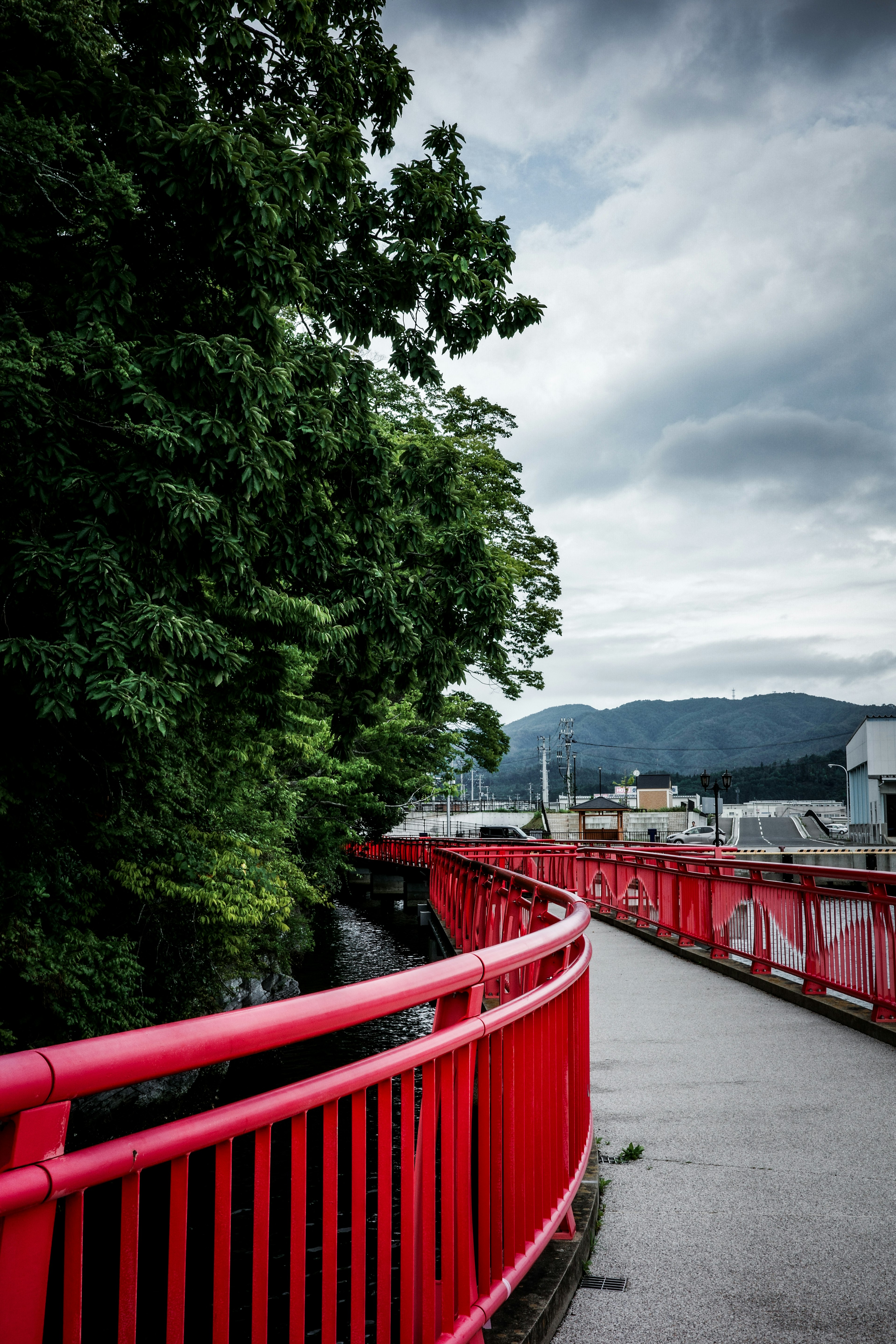 Jembatan melengkung dengan pagar merah dikelilingi oleh pepohonan dan langit mendung