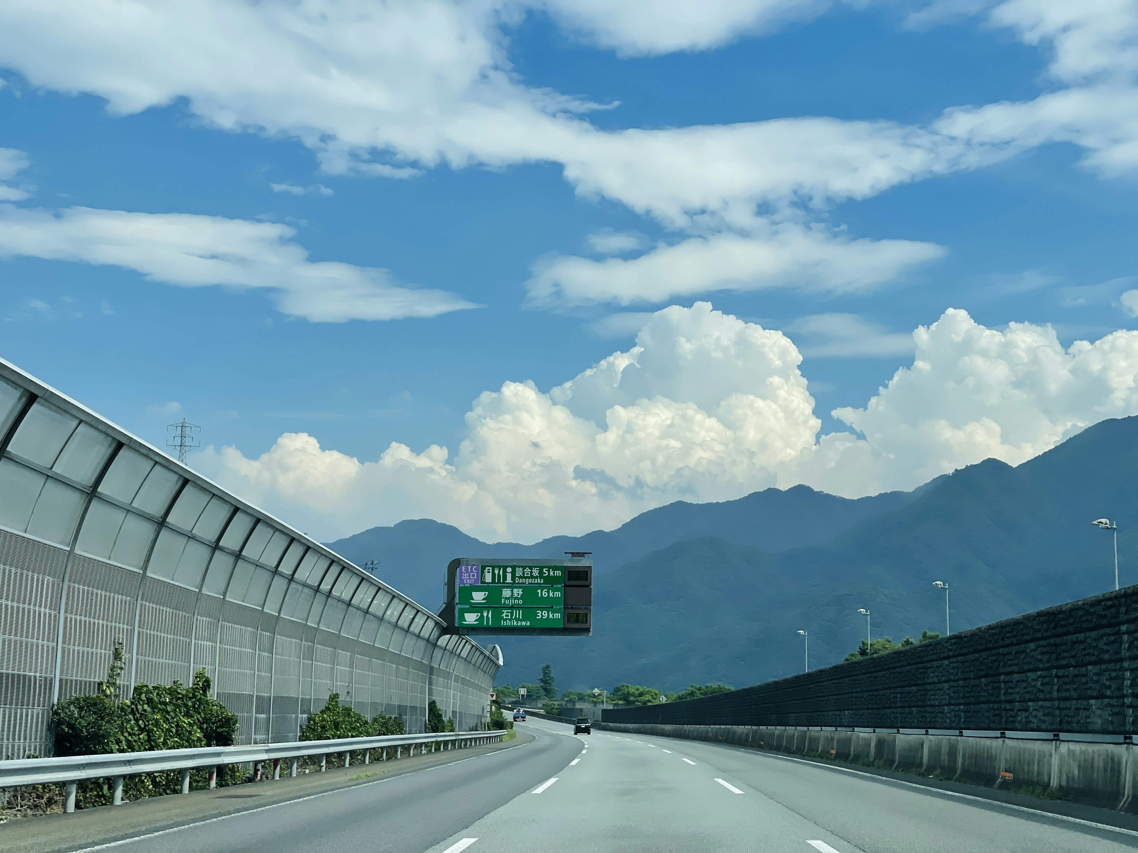 青空と白い雲が広がる山道の風景 高速道路の標識と周囲の緑が特徴