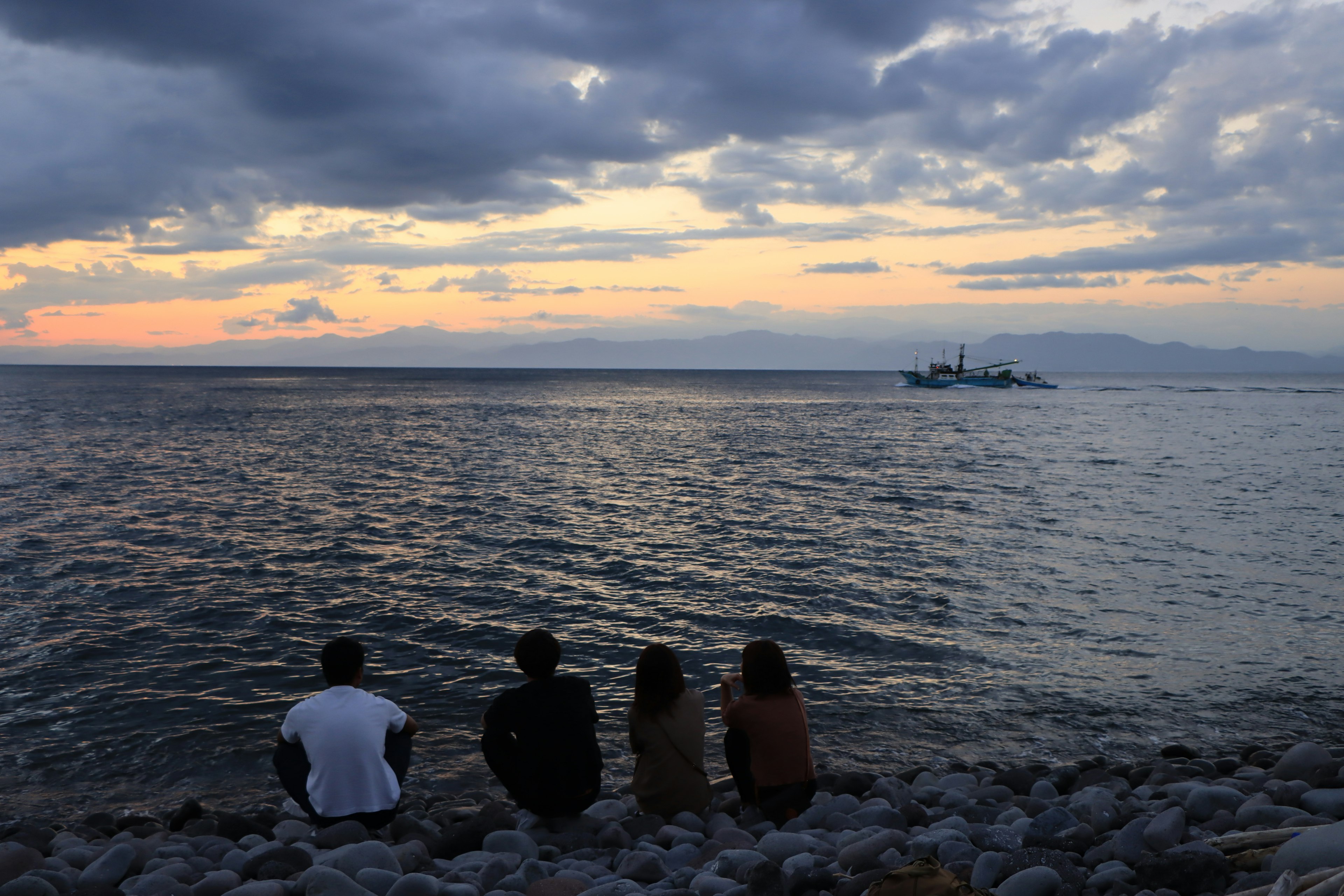 Silueta de cuatro personas sentadas junto al mar con un atardecer de fondo