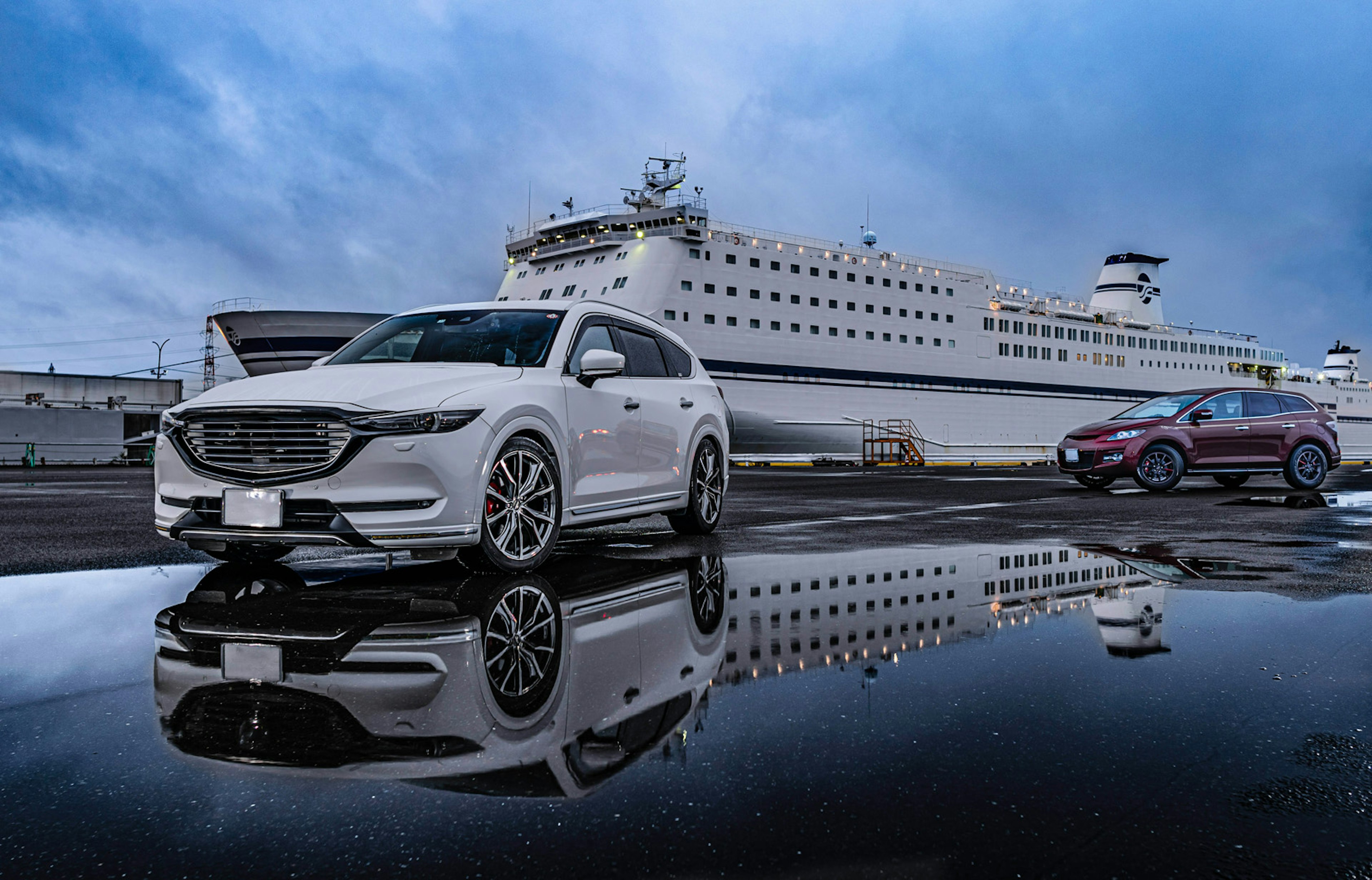 A white car and a red car parked in front of a ship at the harbor with reflections in a puddle