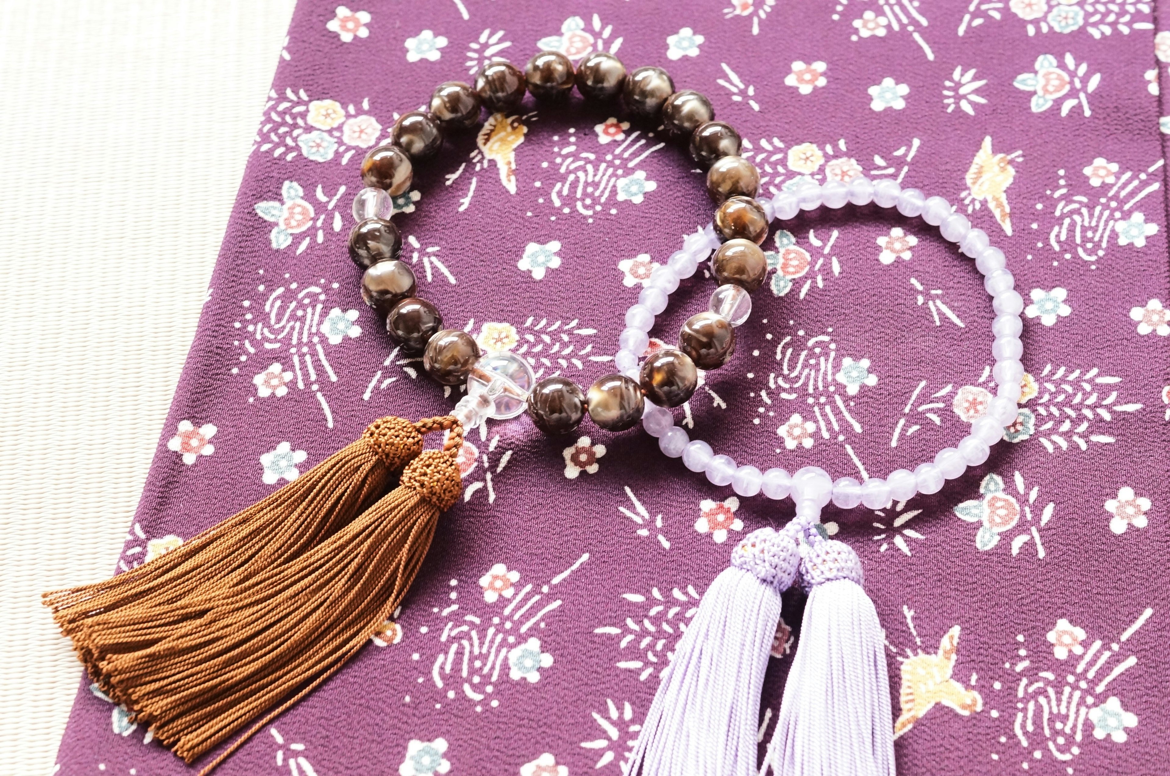 Two bracelets on a purple floral fabric one with brown tassels and the other with white tassels