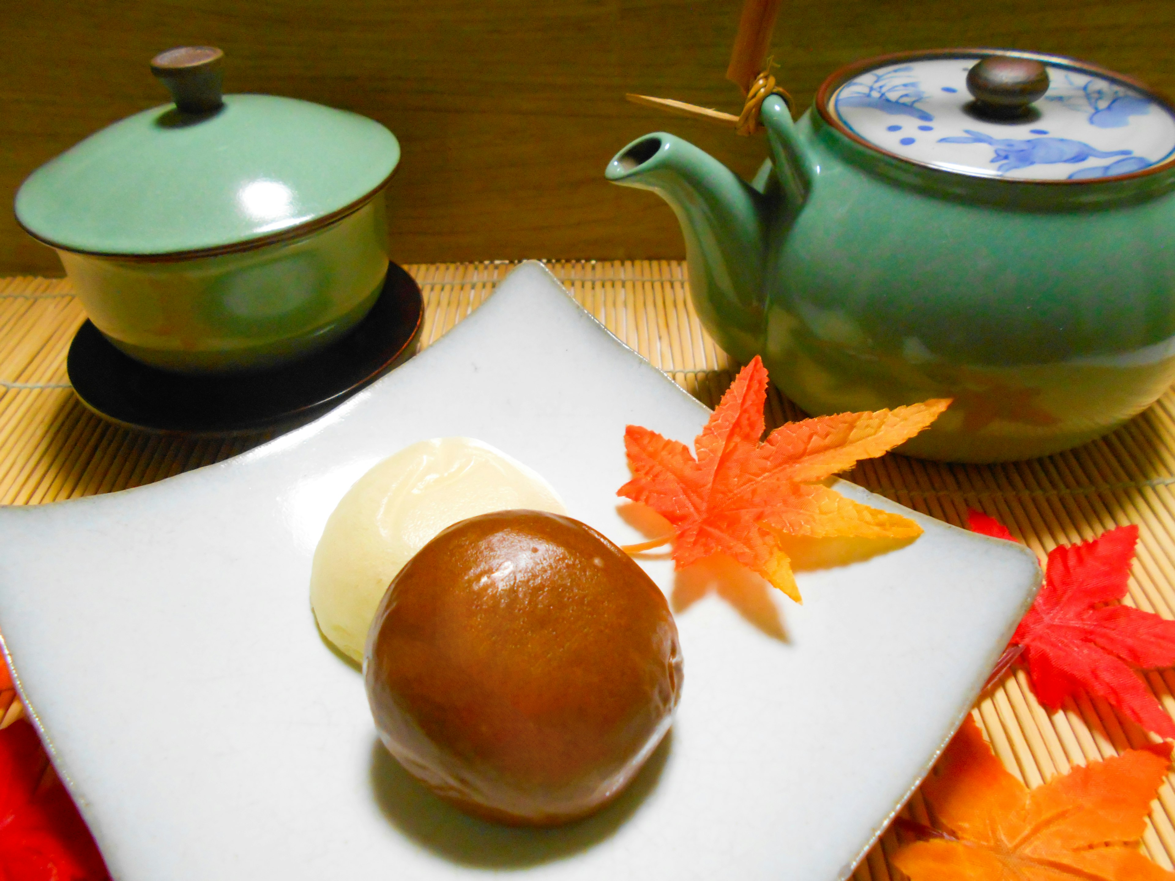 A green teapot and bowls beside a plate with autumn leaves and Japanese sweets