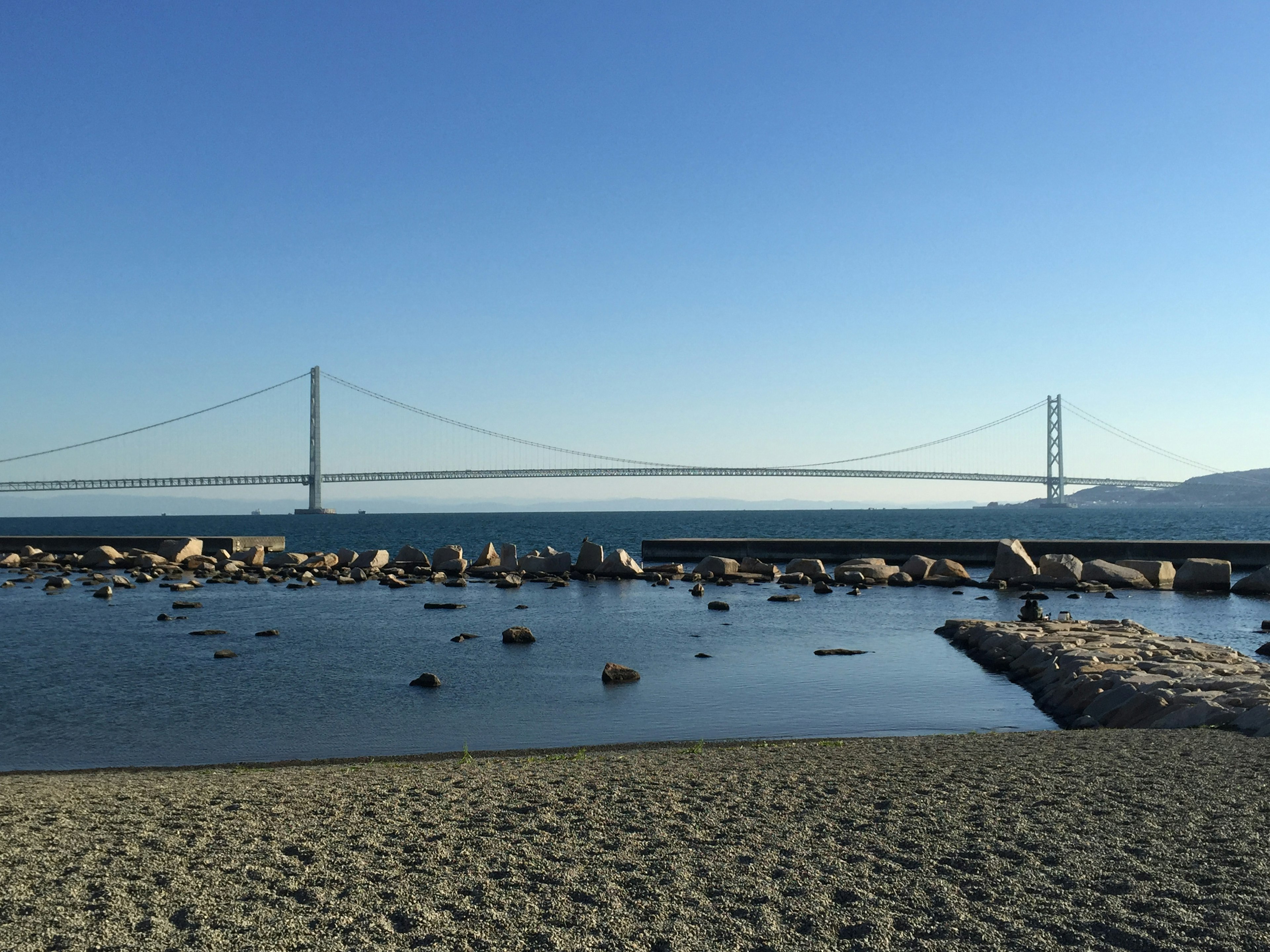 Pantai berpasir dengan laut tenang di bawah langit biru Jembatan Akashi Kaikyō di kejauhan