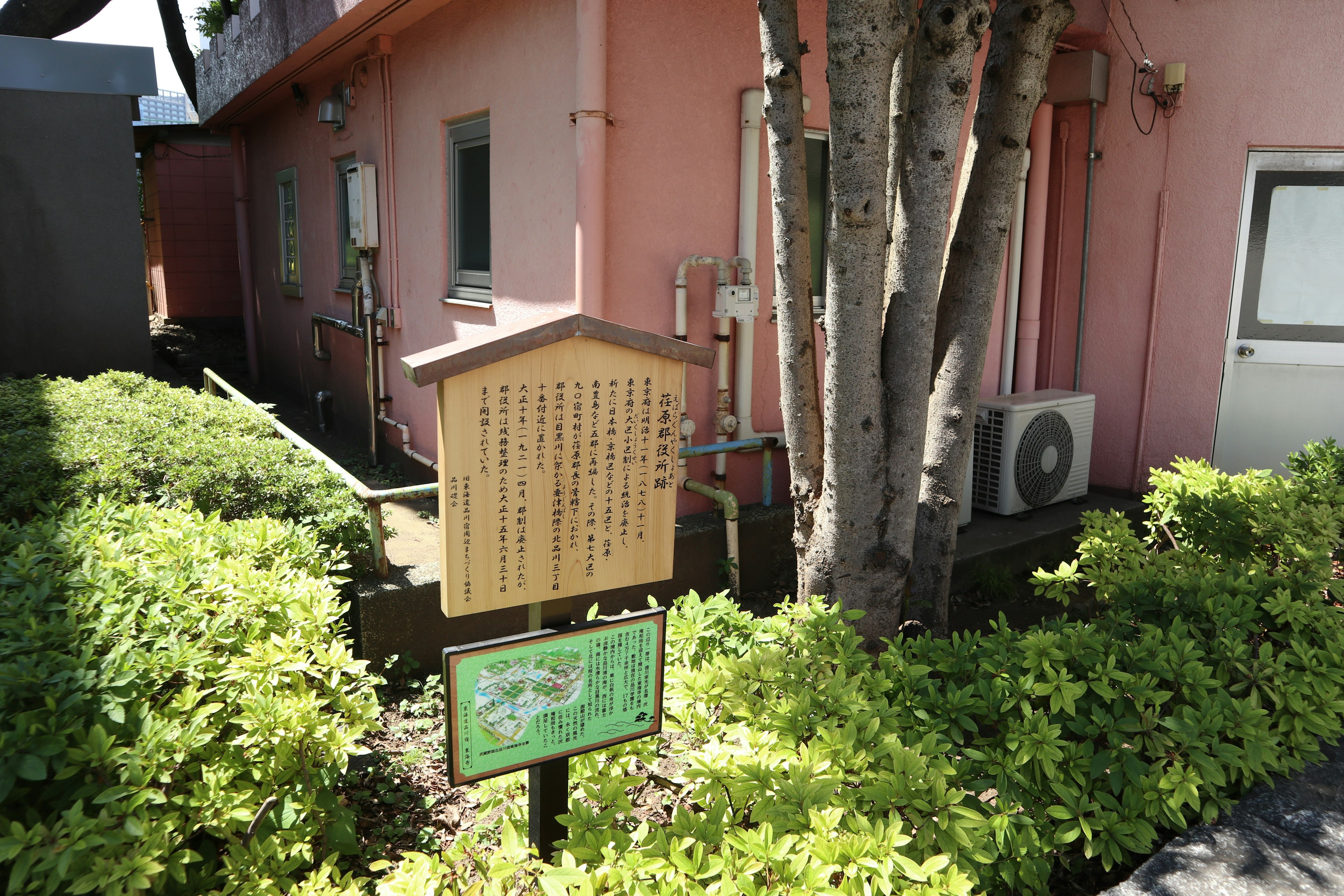 Signboard standing between green plants and trees next to a pink building