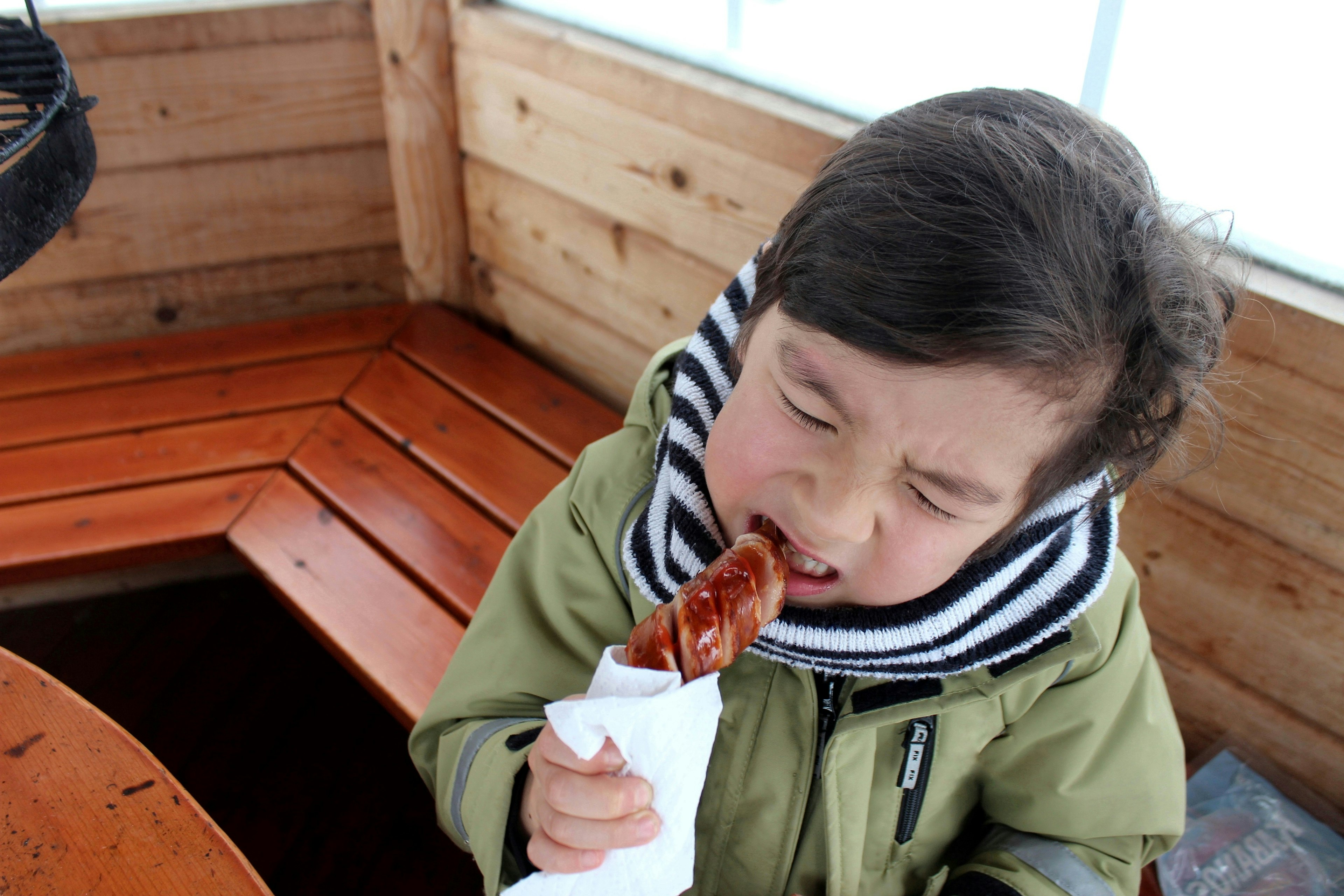 Kind genießt eine Wurst in einer gemütlichen Holzumgebung mit warmer Kleidung