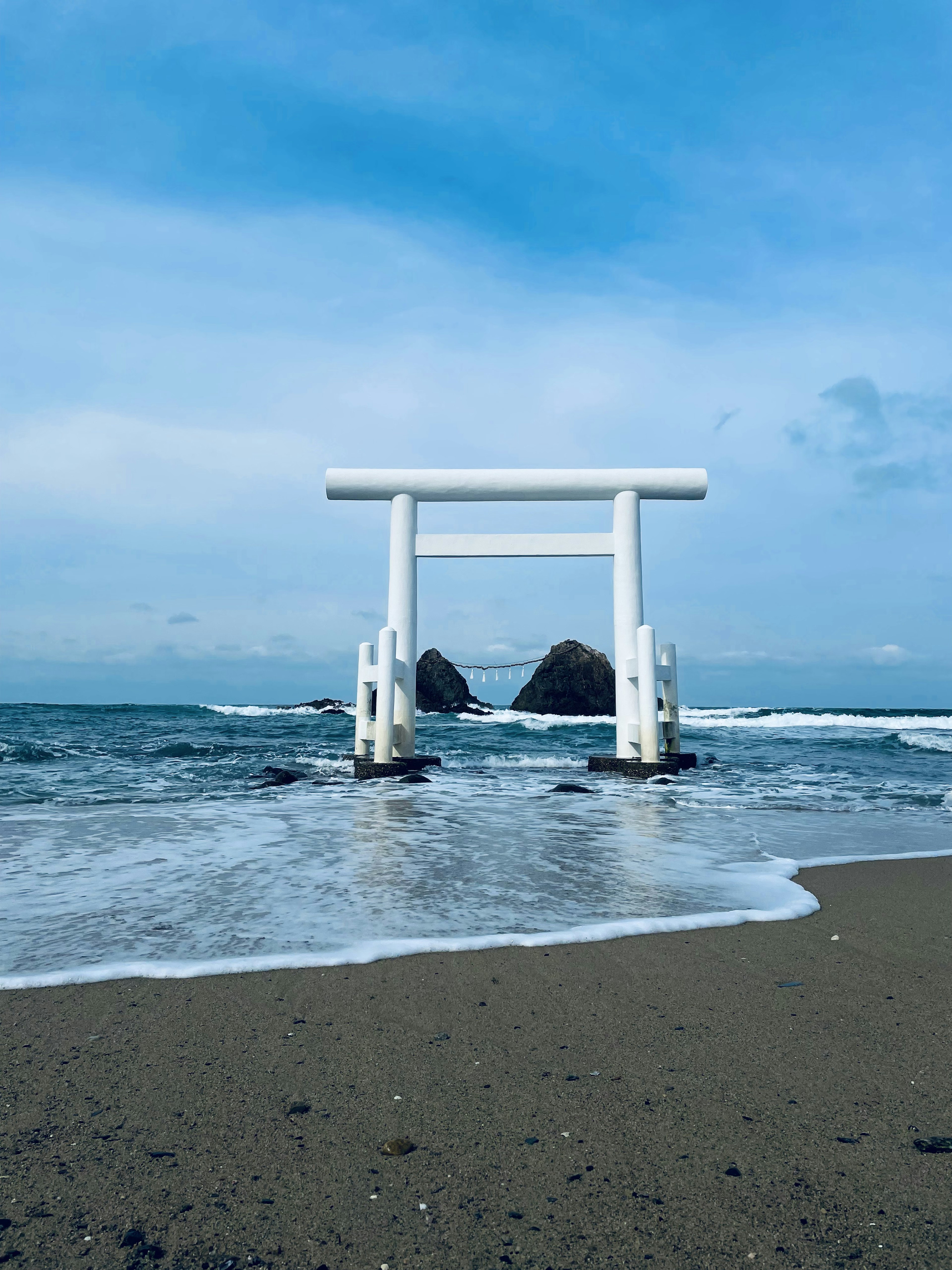 A white torii gate stands in the ocean with rocky islands in the background