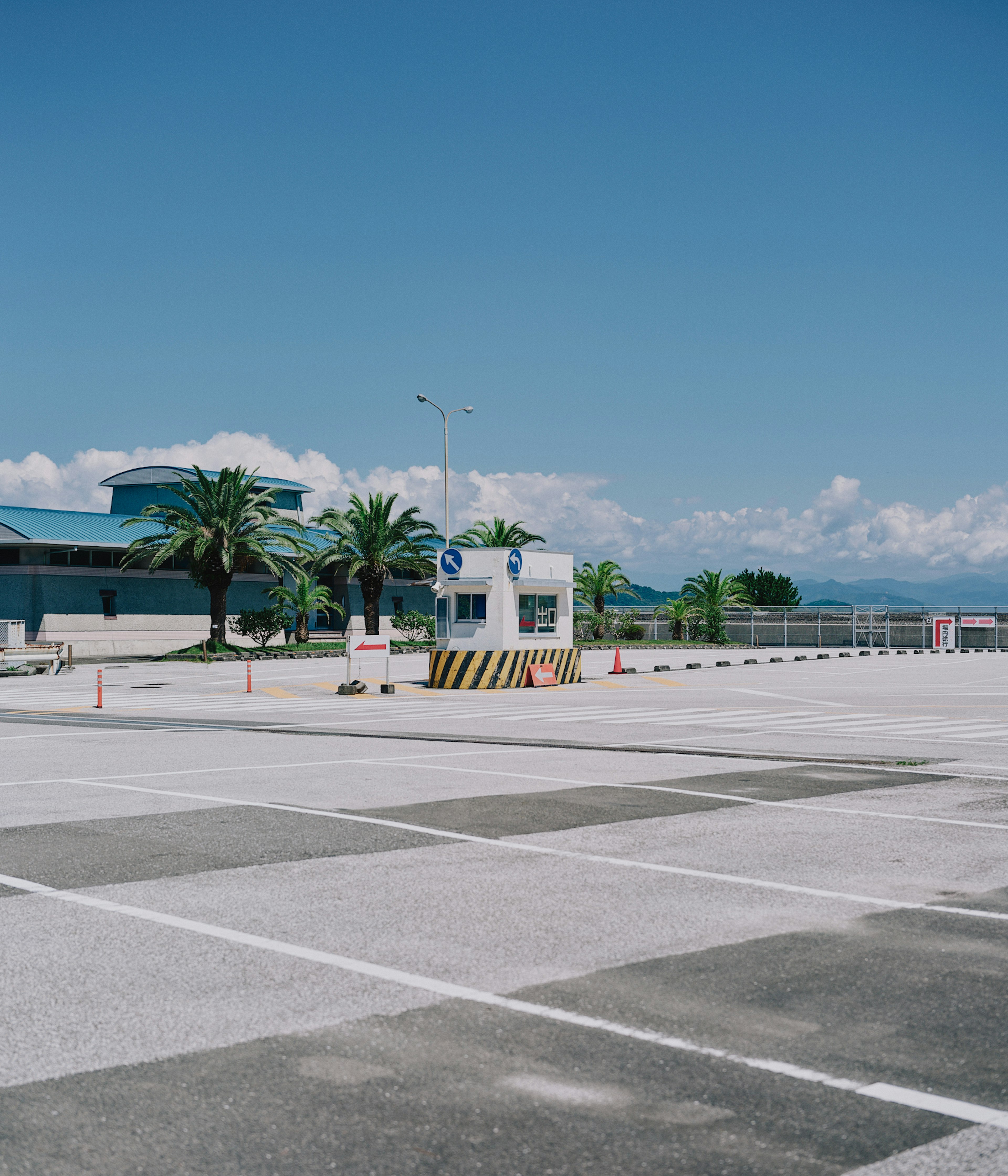 Tempat parkir bandara dengan pos jaga dan pohon palem di bawah langit biru cerah