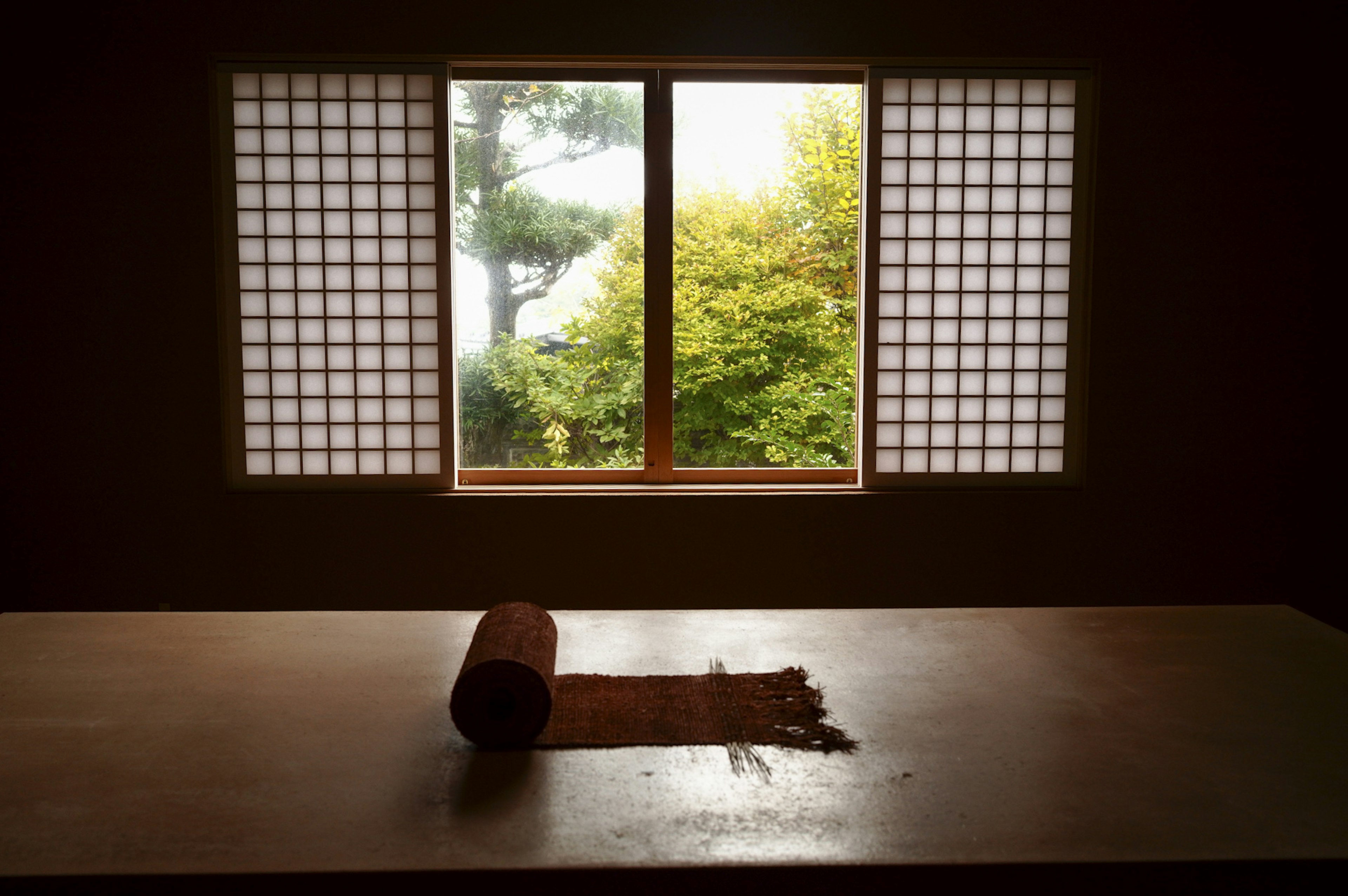 Habitación japonesa tradicional con ventana mostrando un paisaje verde y una alfombra enrollada en la mesa