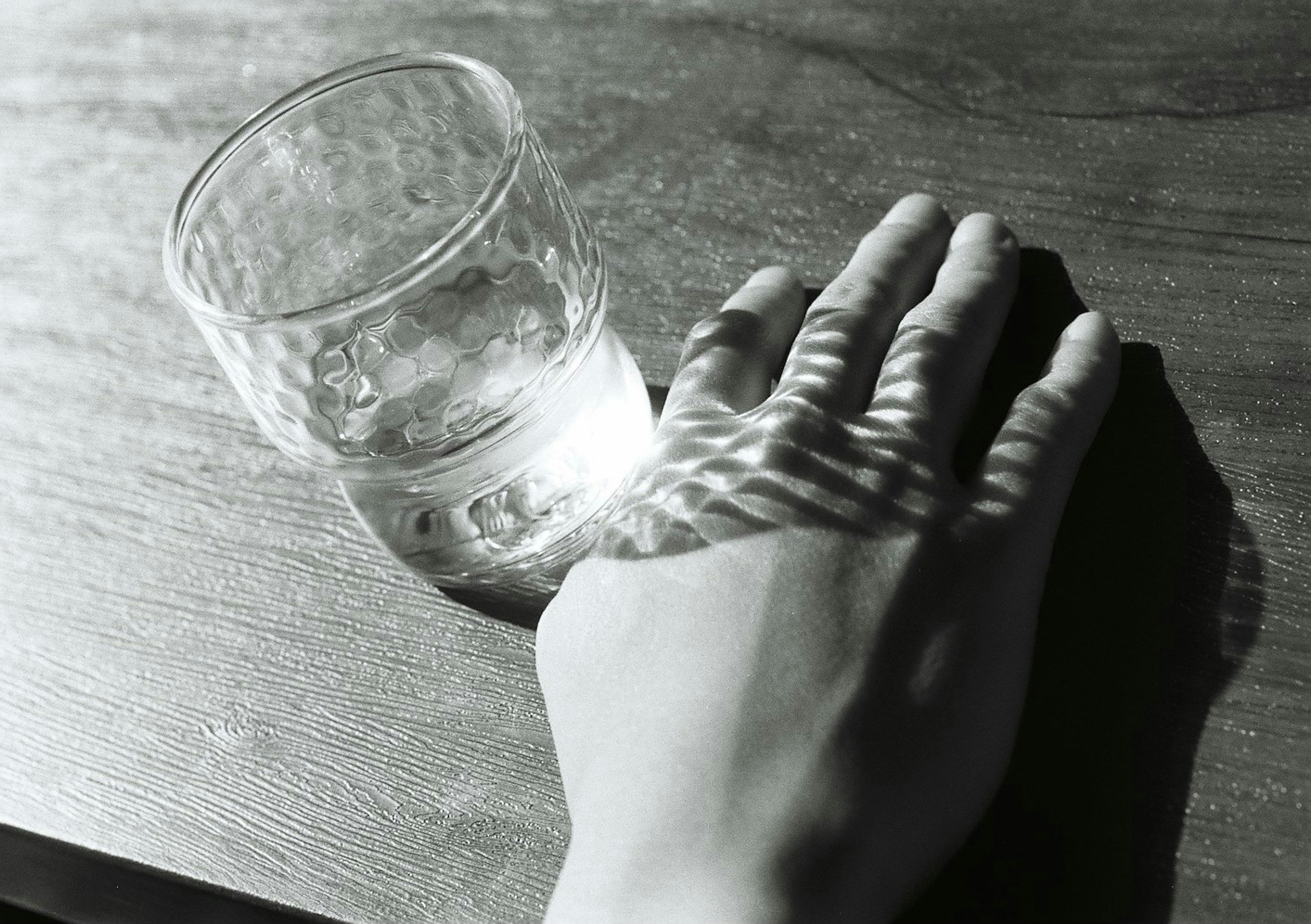 Eine Hand, die auf einem Tisch neben einem Wasserglas in Schwarzweiß ruht