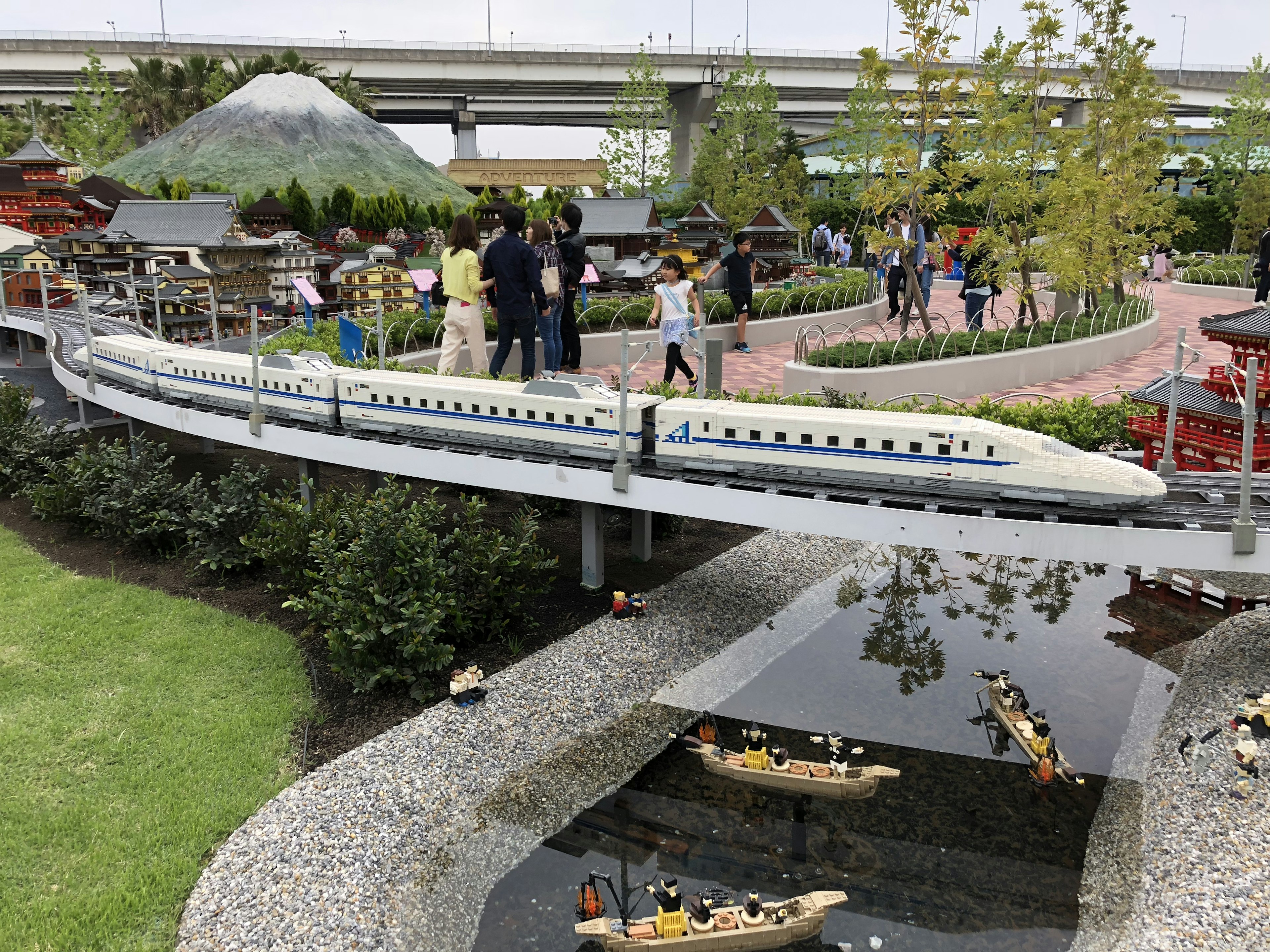 A miniature Shinkansen train crossing a bridge in a park with visitors