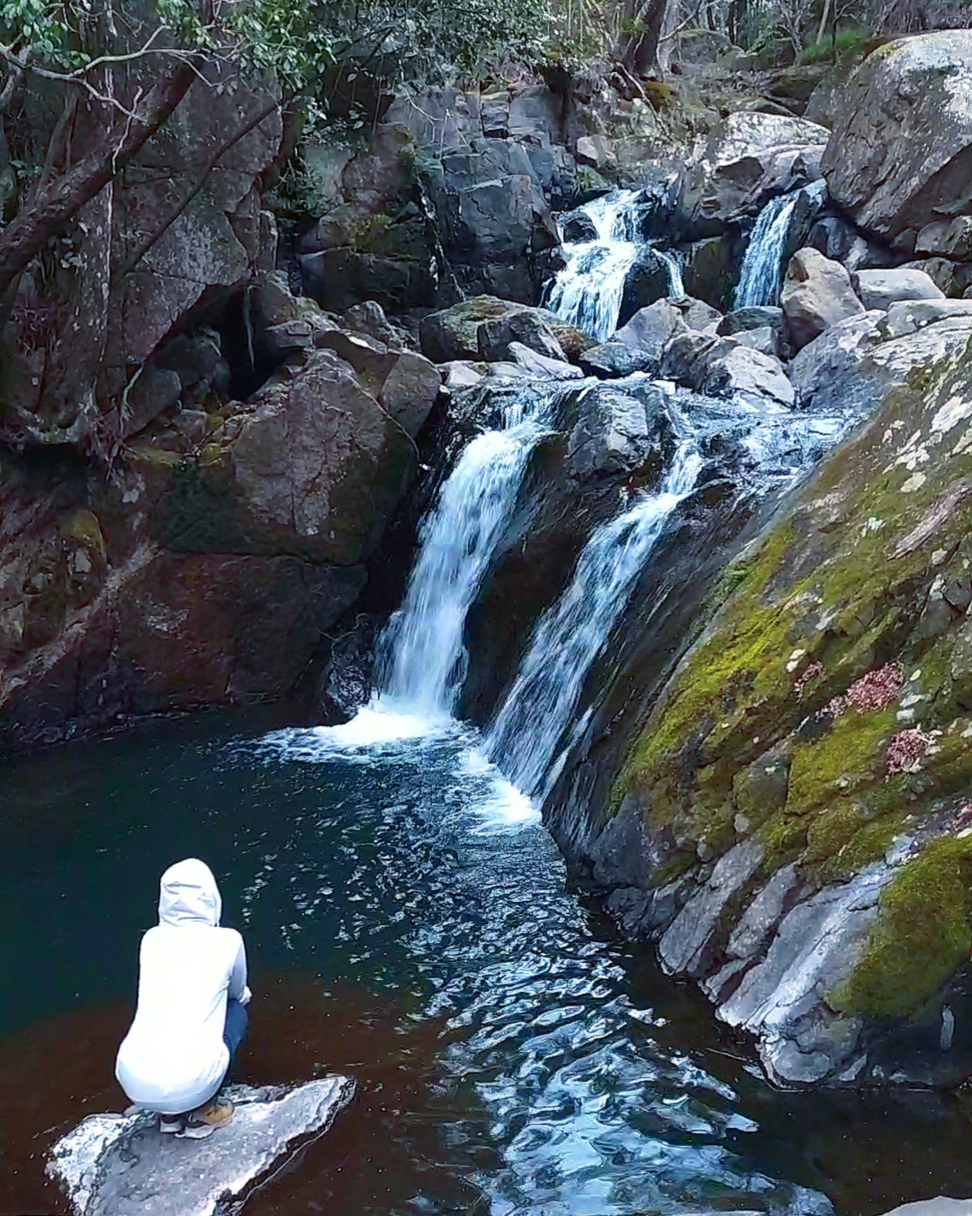 Eine Person in einem weißen Outfit sitzt auf einem Felsen neben einem Wasserfall