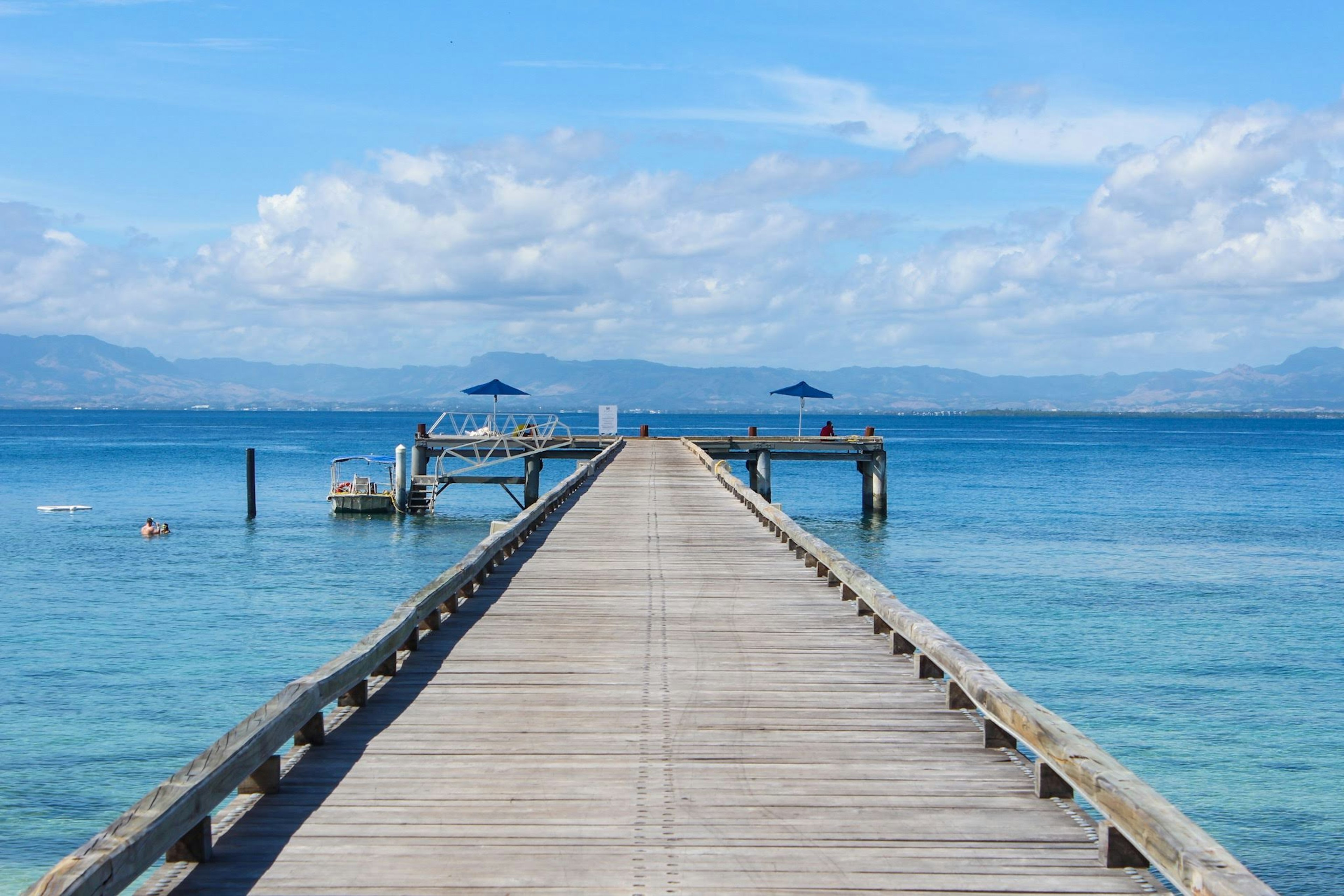 Dermaga kayu yang membentang ke laut biru di bawah langit cerah dengan awan putih