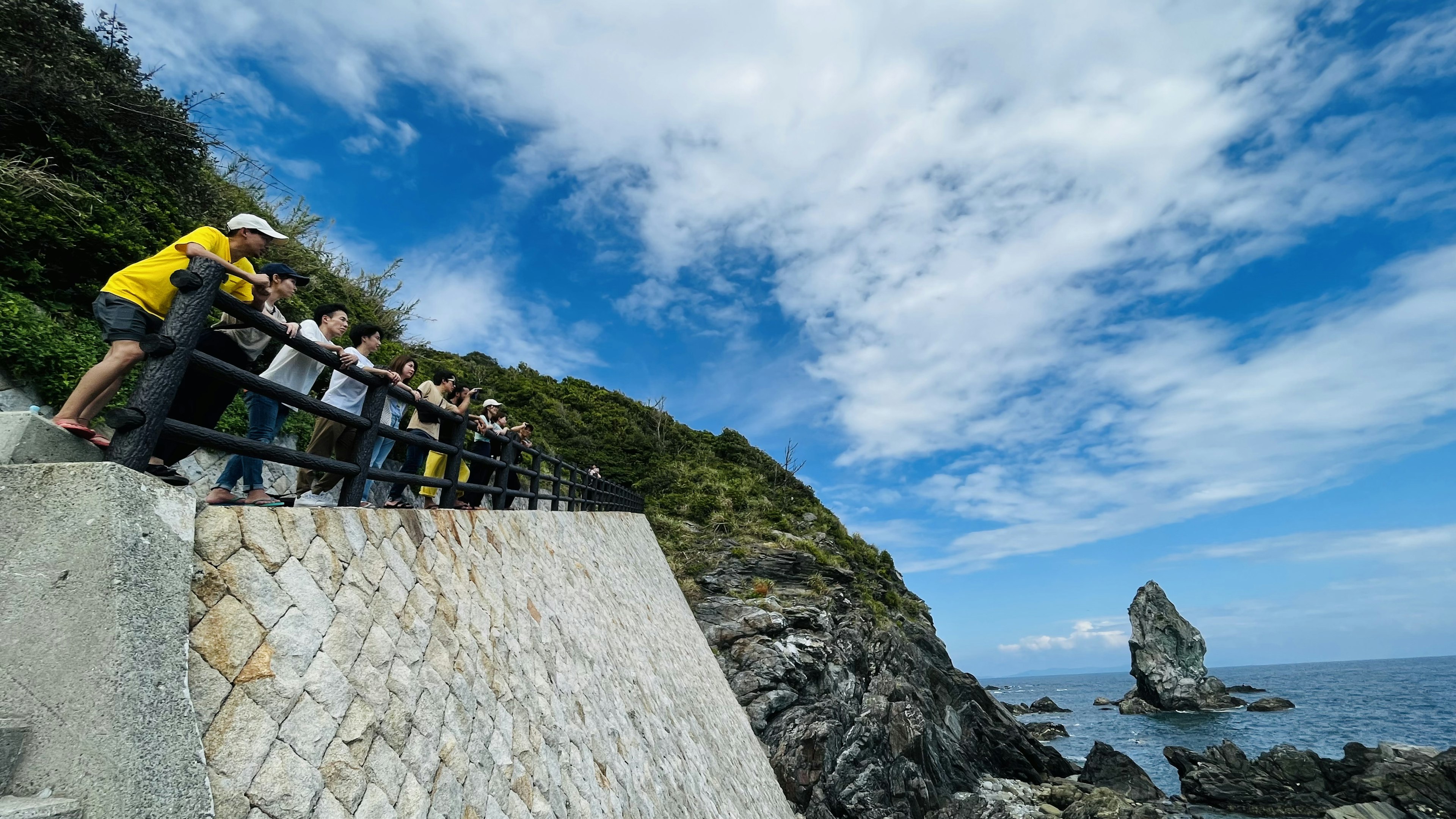 Personas mirando la costa con una roca prominente bajo un cielo azul