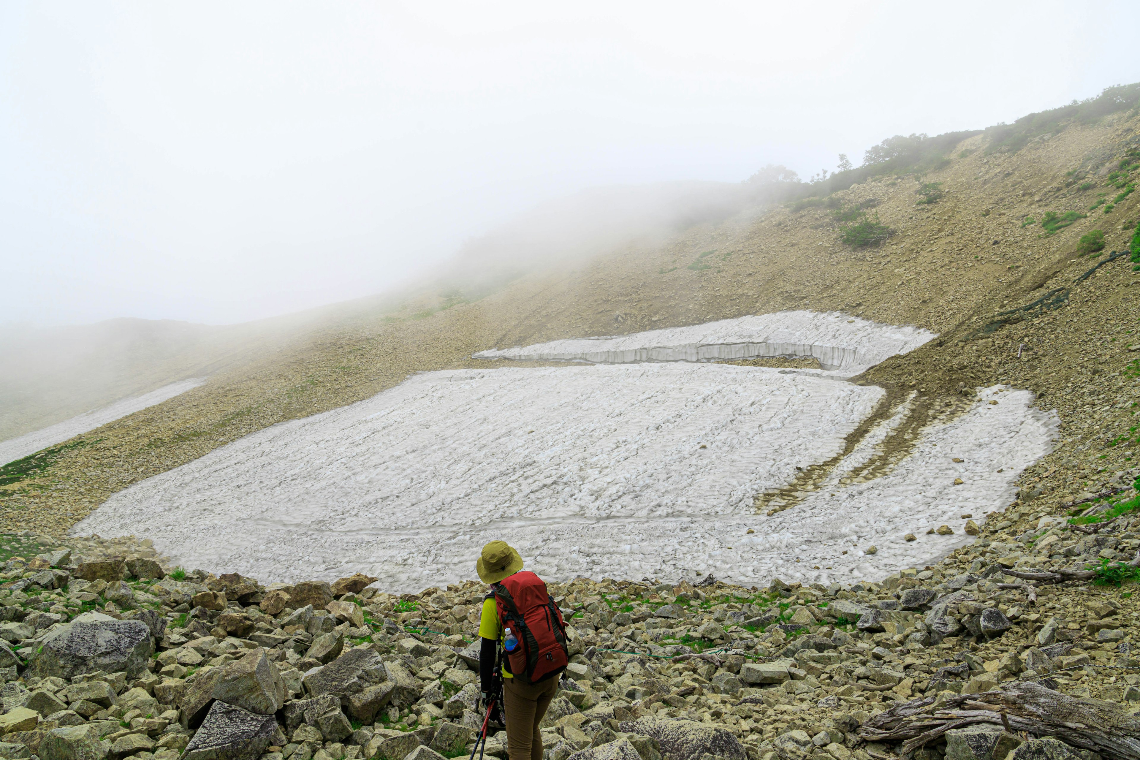 Hiker observing a snow patch on a mountain slope in fog