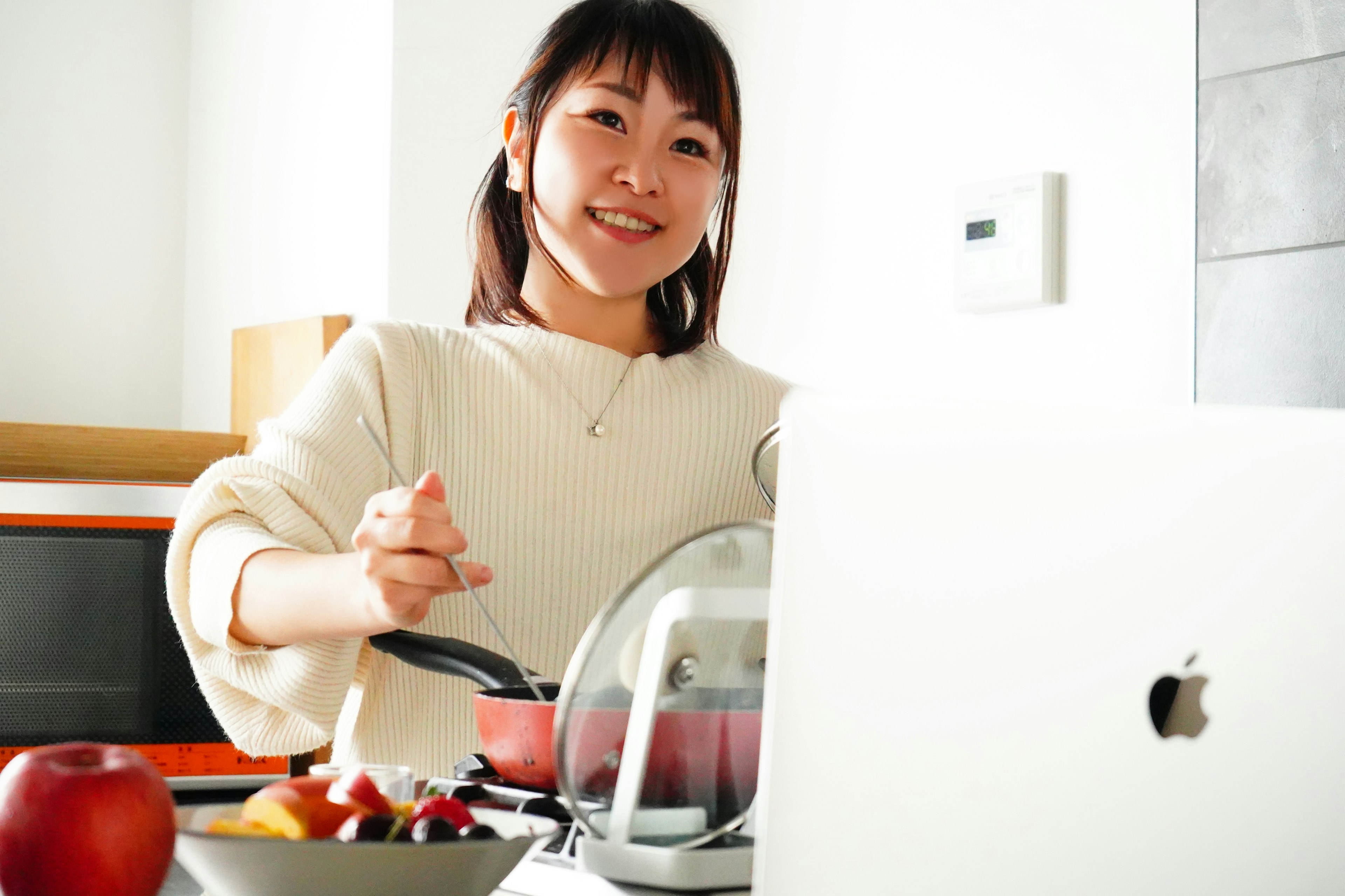 Mujer cocinando en una cocina sonriendo mientras revuelve una olla con manzanas y un tazón de frutas cerca