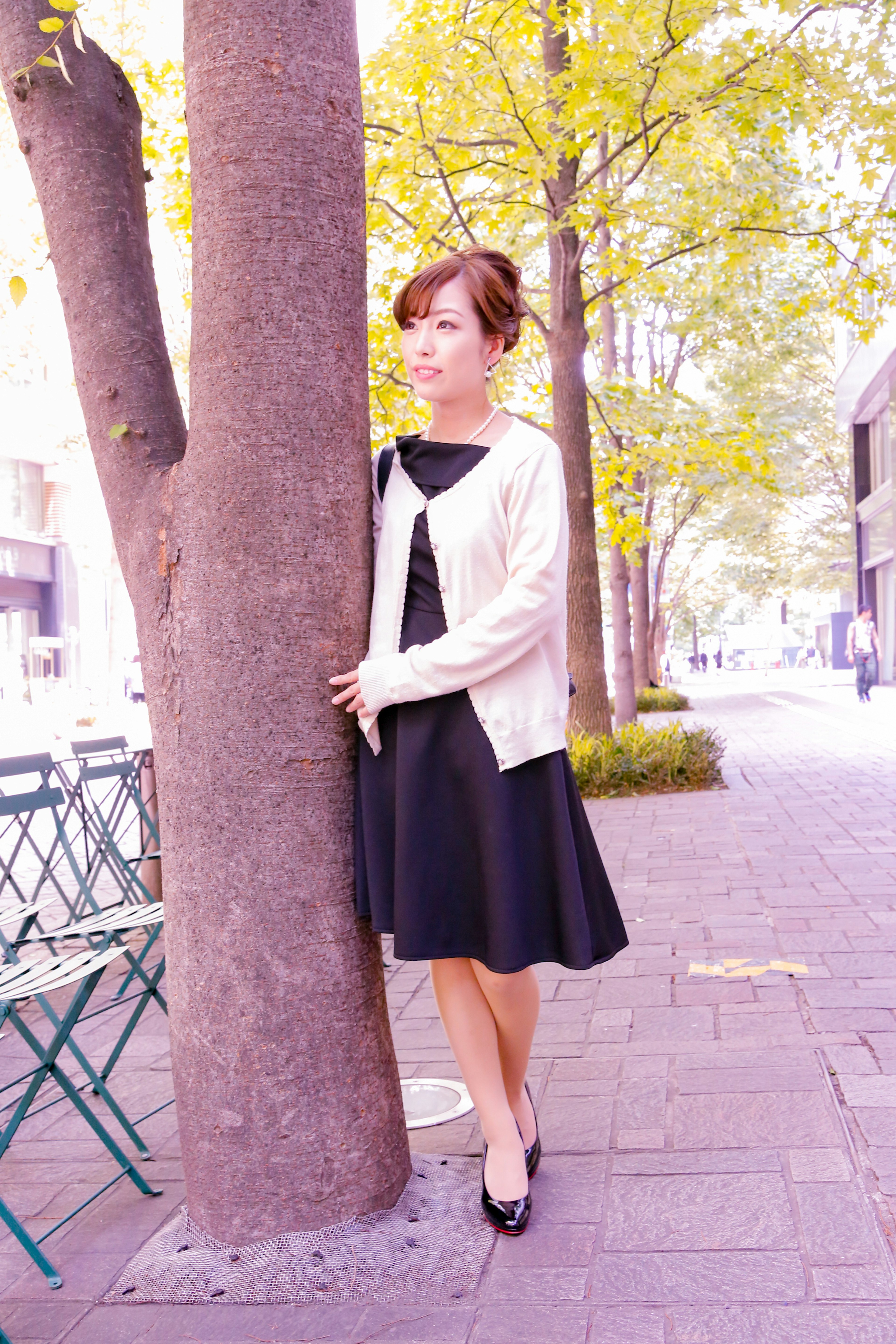 Woman standing by a tree in a park wearing a black dress and white cardigan with yellow-leaved trees in the background