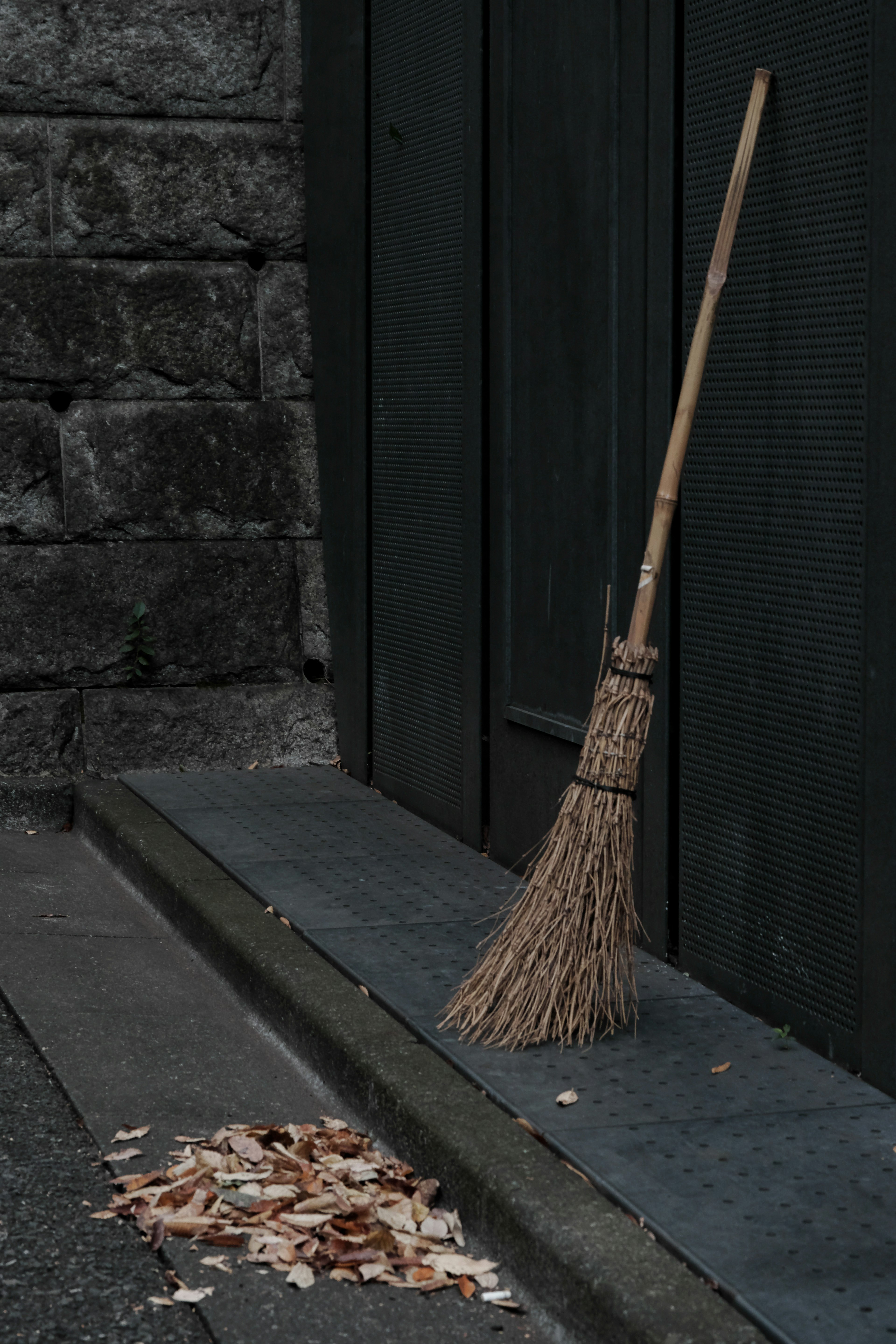 A broom leaning against a dark green wall with scattered leaves on the ground