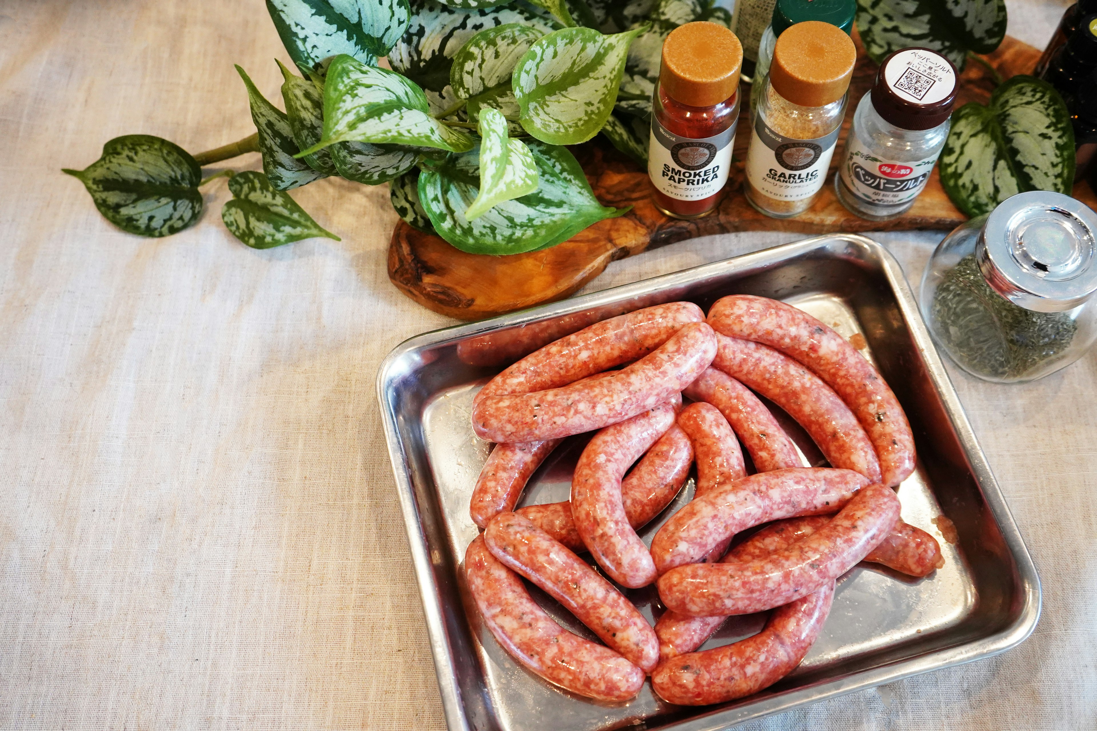 Raw sausages arranged on a metal tray with spice jars and green plants in the background