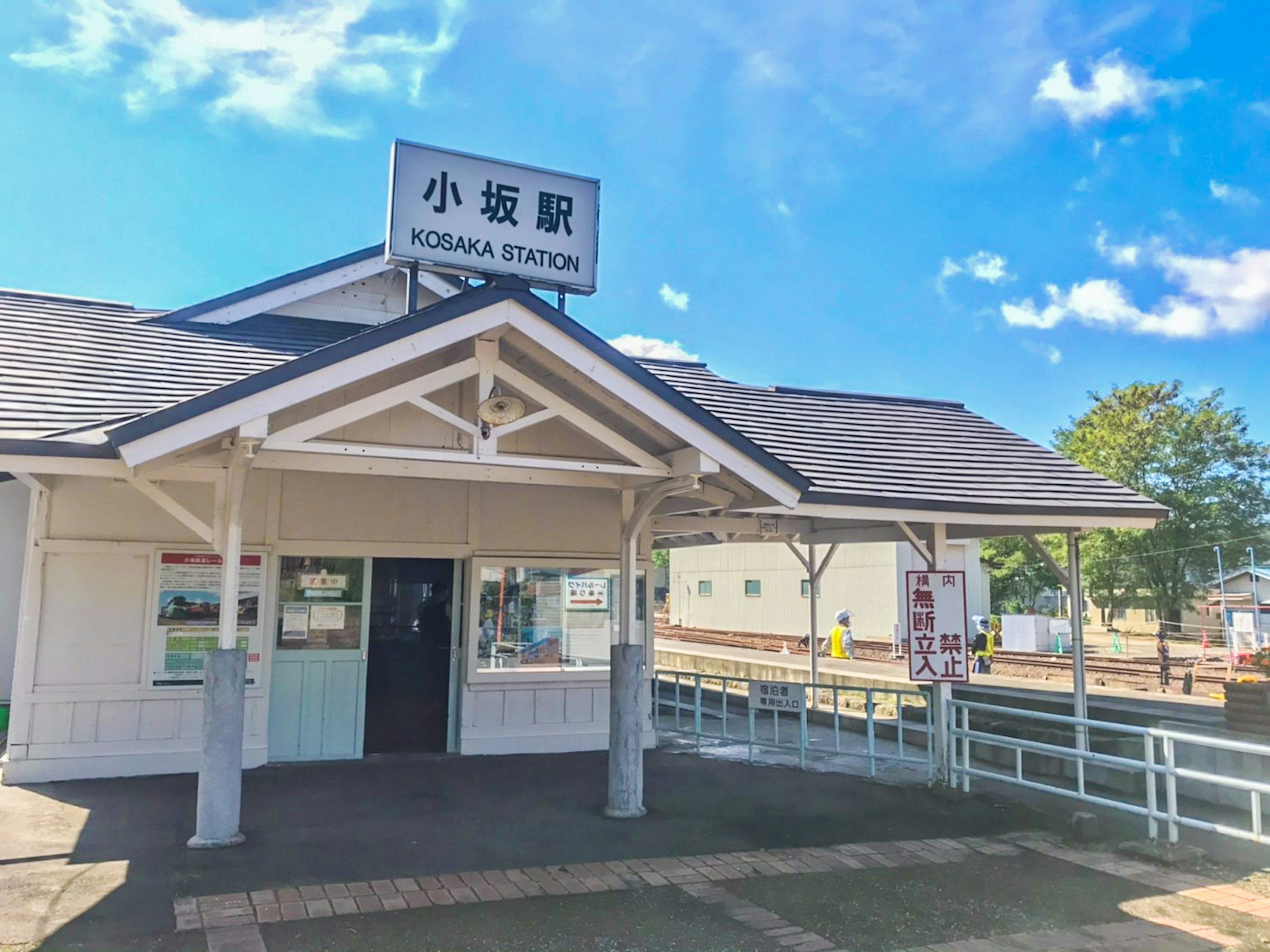 Exterior de la estación de Kosaka, estructura de madera blanca, cielo azul, paisaje verde