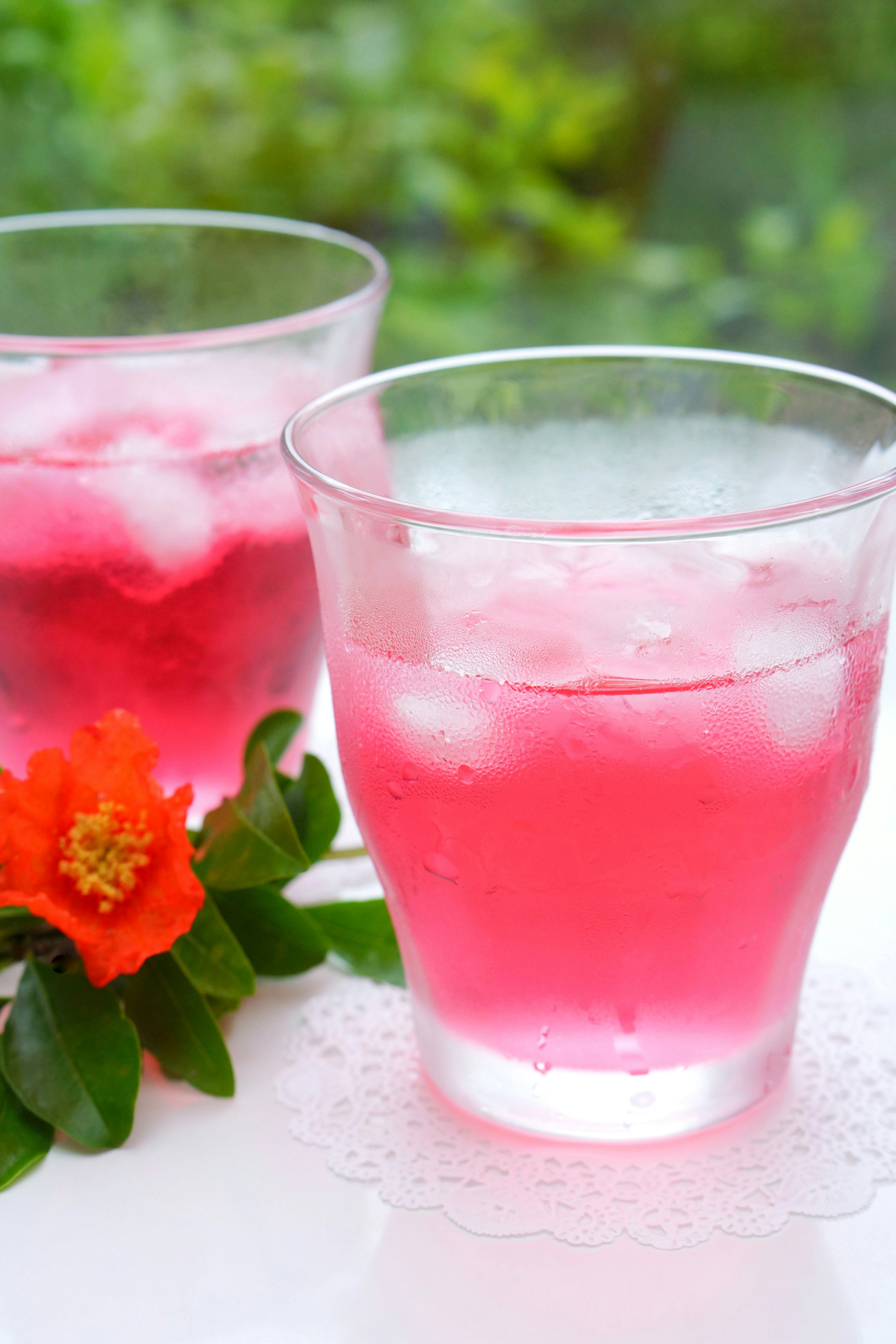 Two clear glasses with pink drink and ice beside a flower