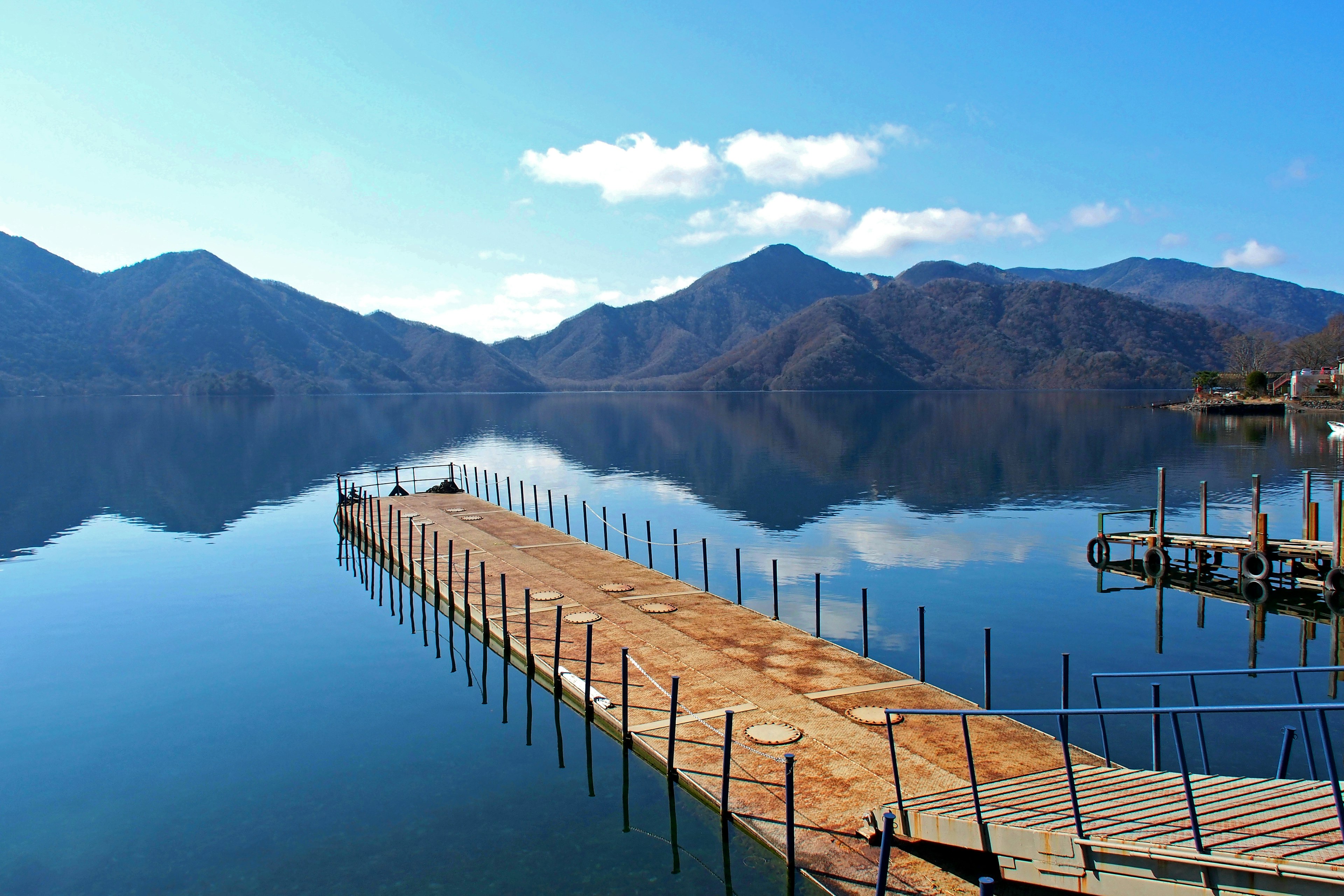 Danau tenang dengan dermaga yang memantulkan gunung dan langit biru