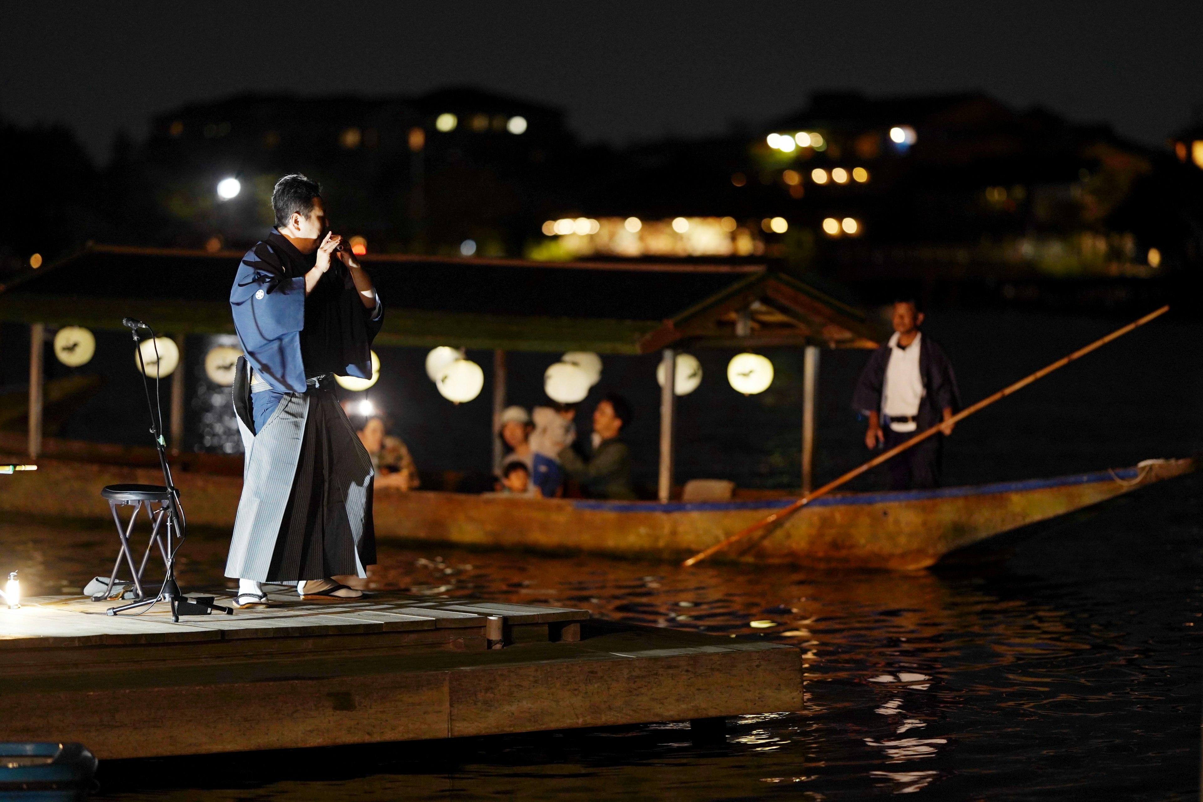 Hombre con vestimenta tradicional cantando junto al agua de noche con un bote al fondo