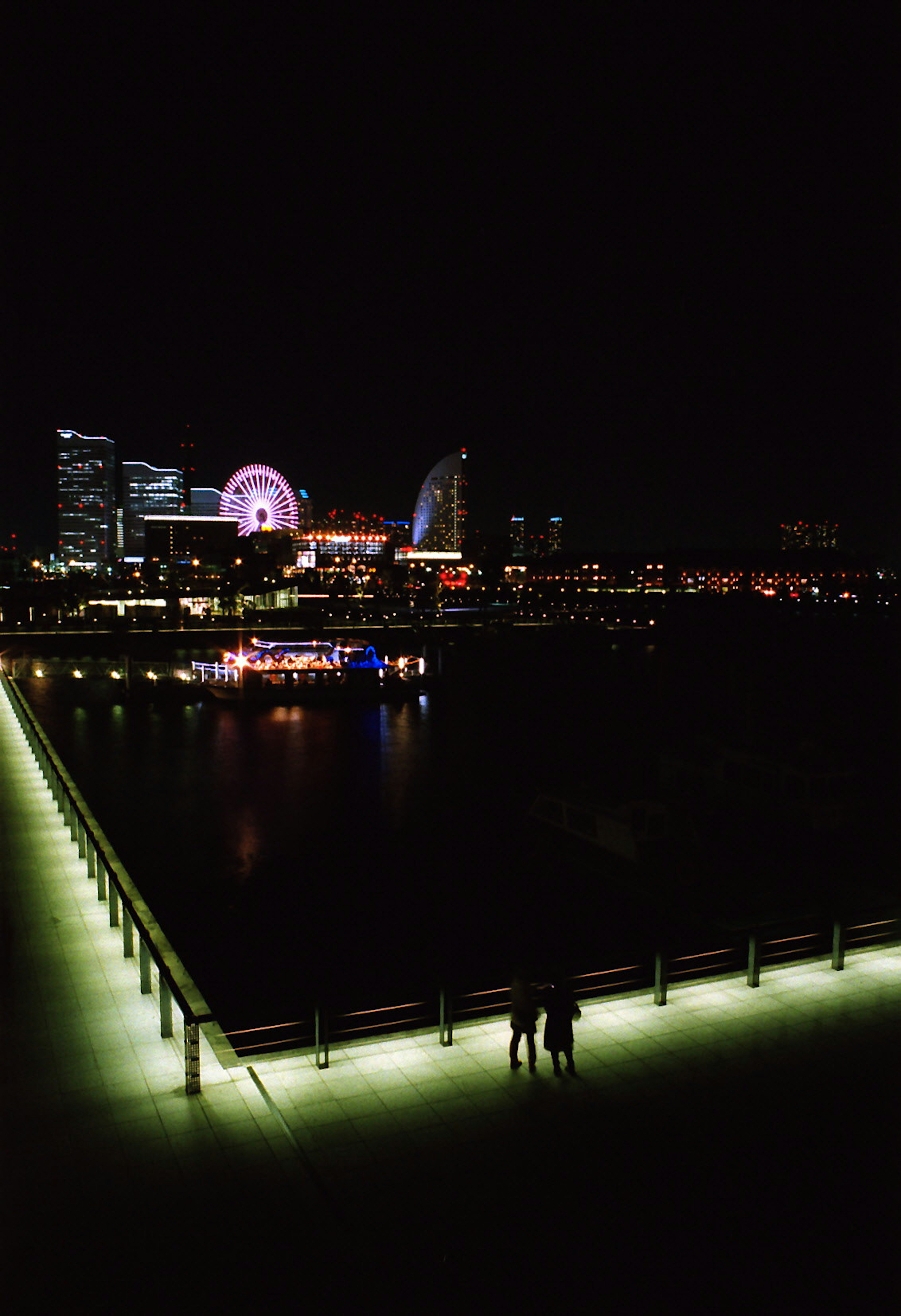 Pareja caminando por un muelle iluminado con el horizonte de la ciudad de noche