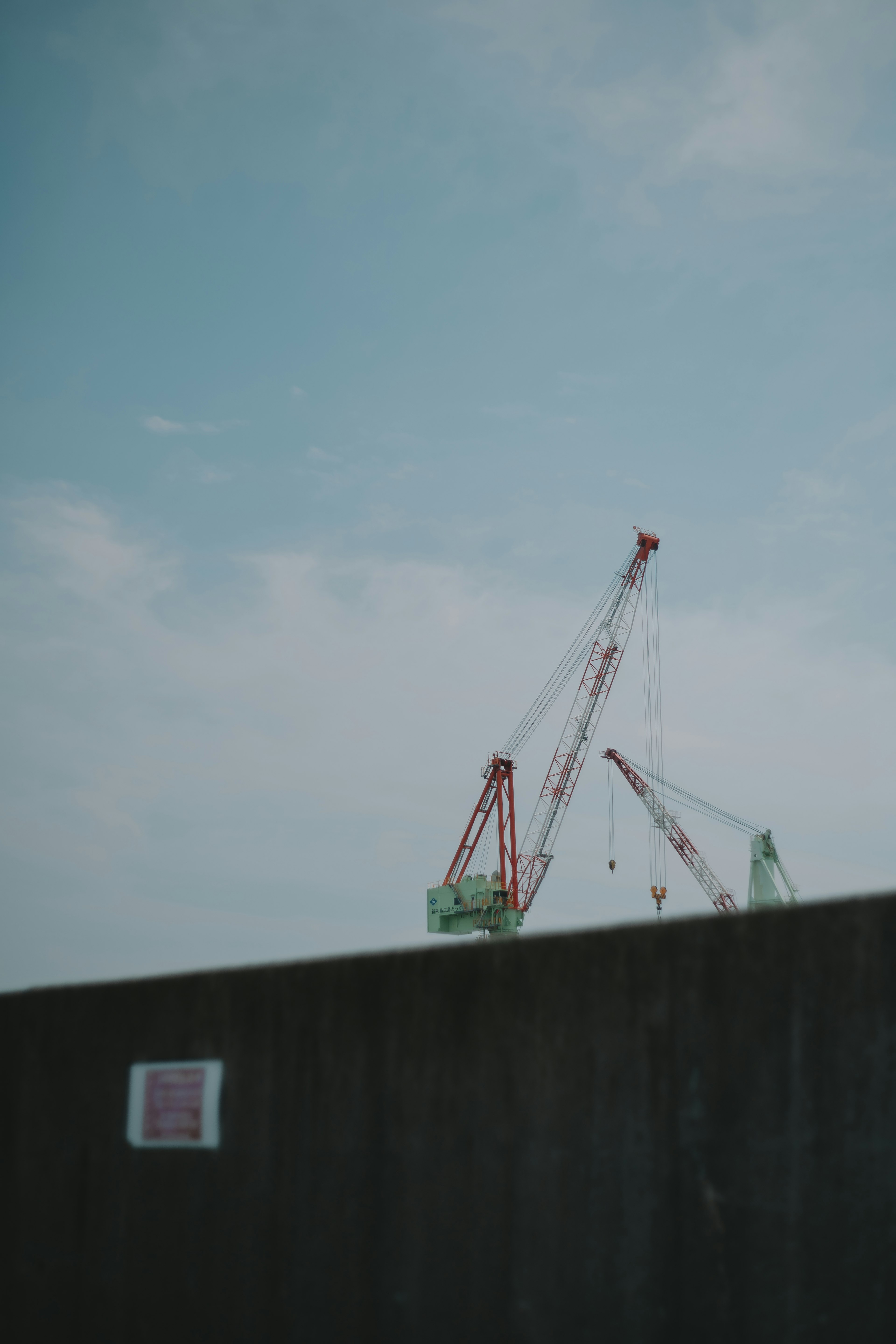 Two cranes under a blue sky with a concrete wall in the foreground