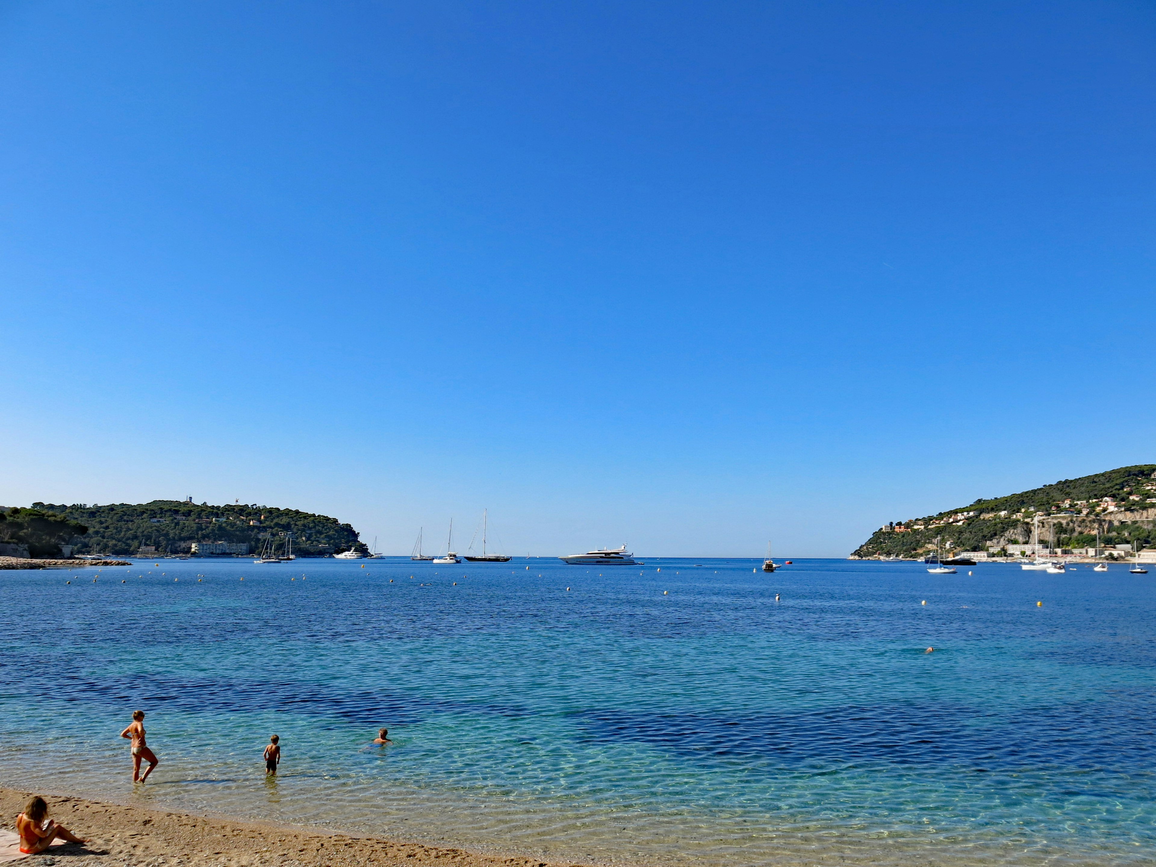 Beach scene with clear blue sea and sky Children playing by the water Boats floating in a calm bay