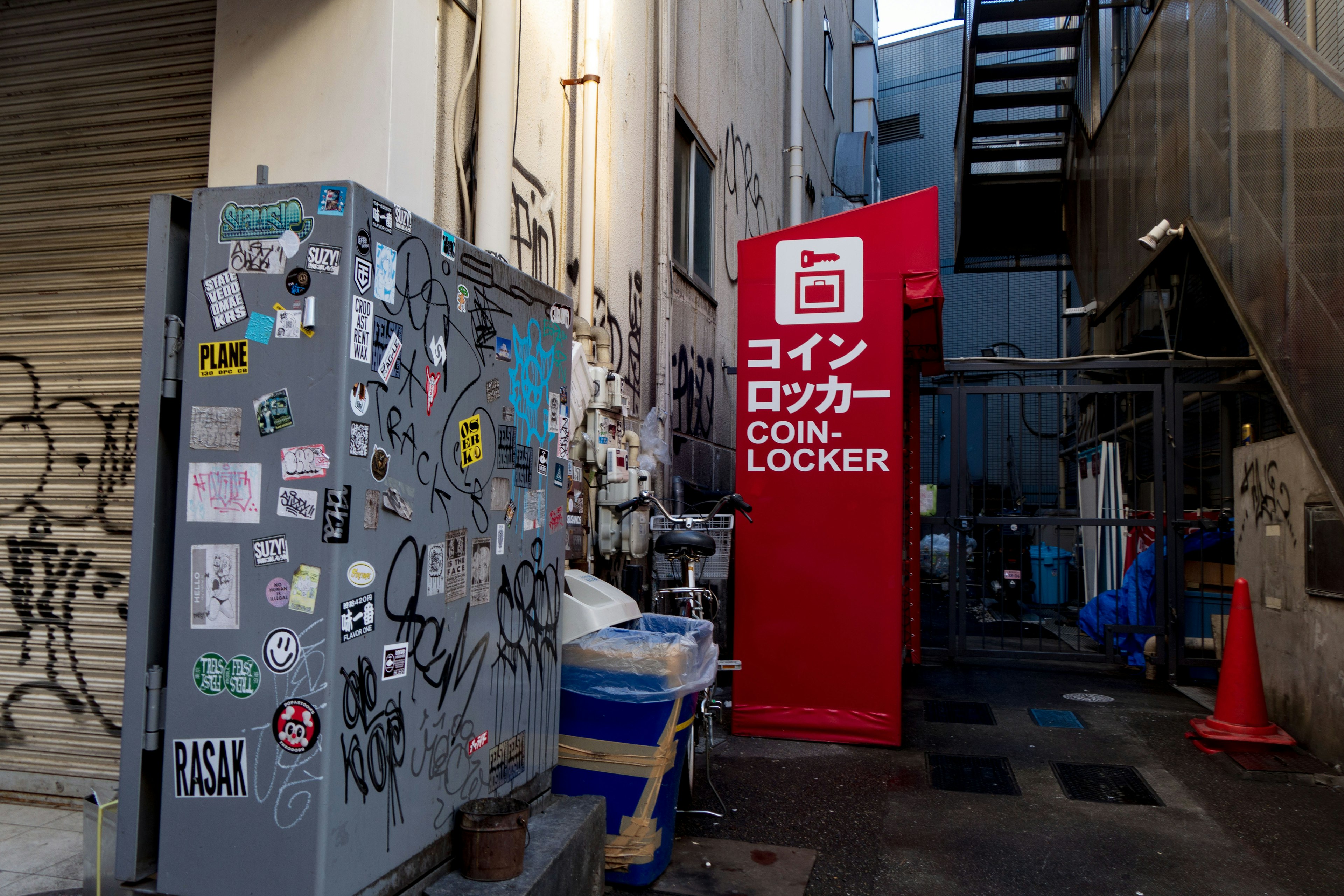 Coin locker in a narrow alley with a sticker-covered utility box