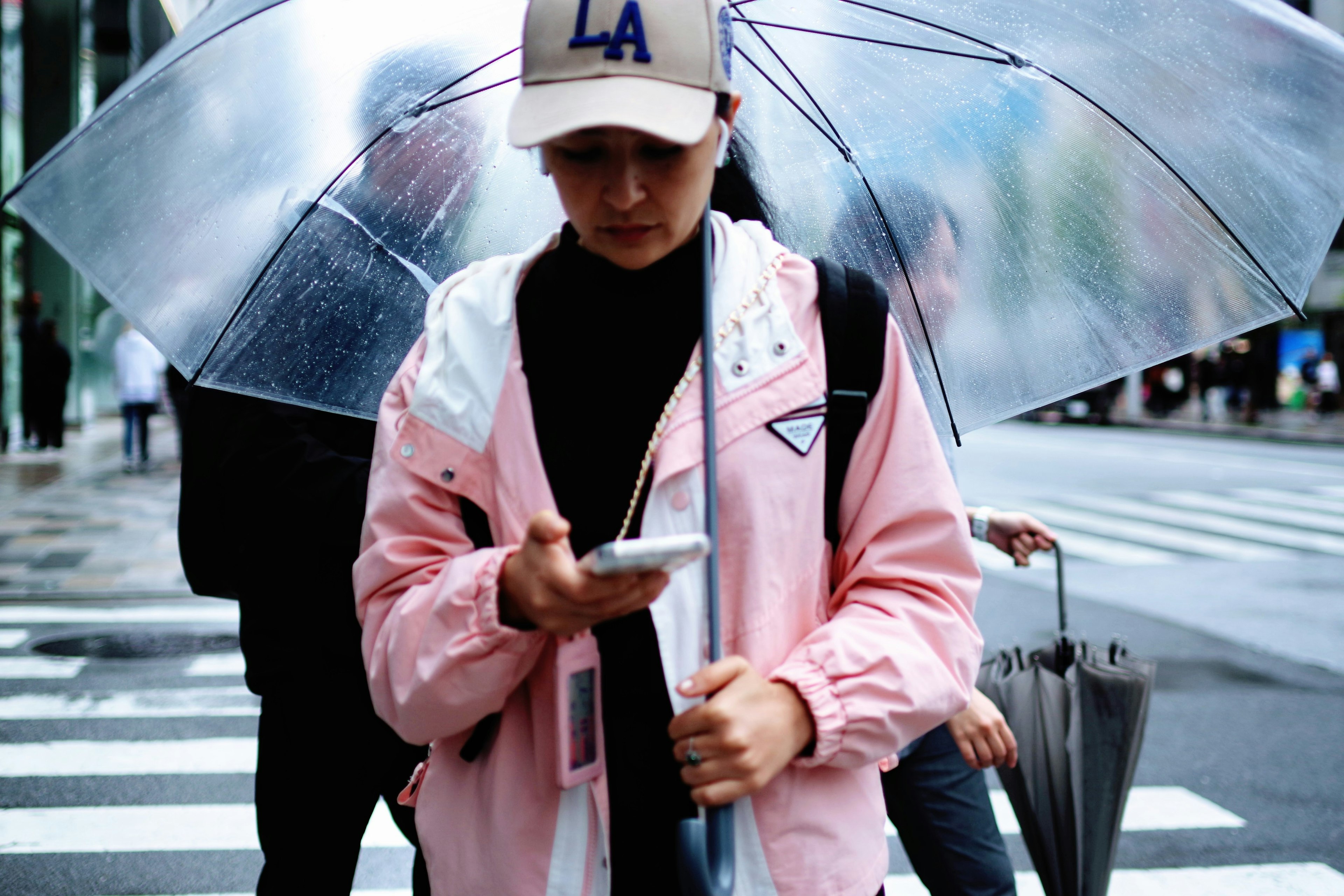 Junger Mensch, der bei Regen ein Smartphone benutzt, trägt eine rosa Jacke und eine LA-Cap, hält einen transparenten Regenschirm