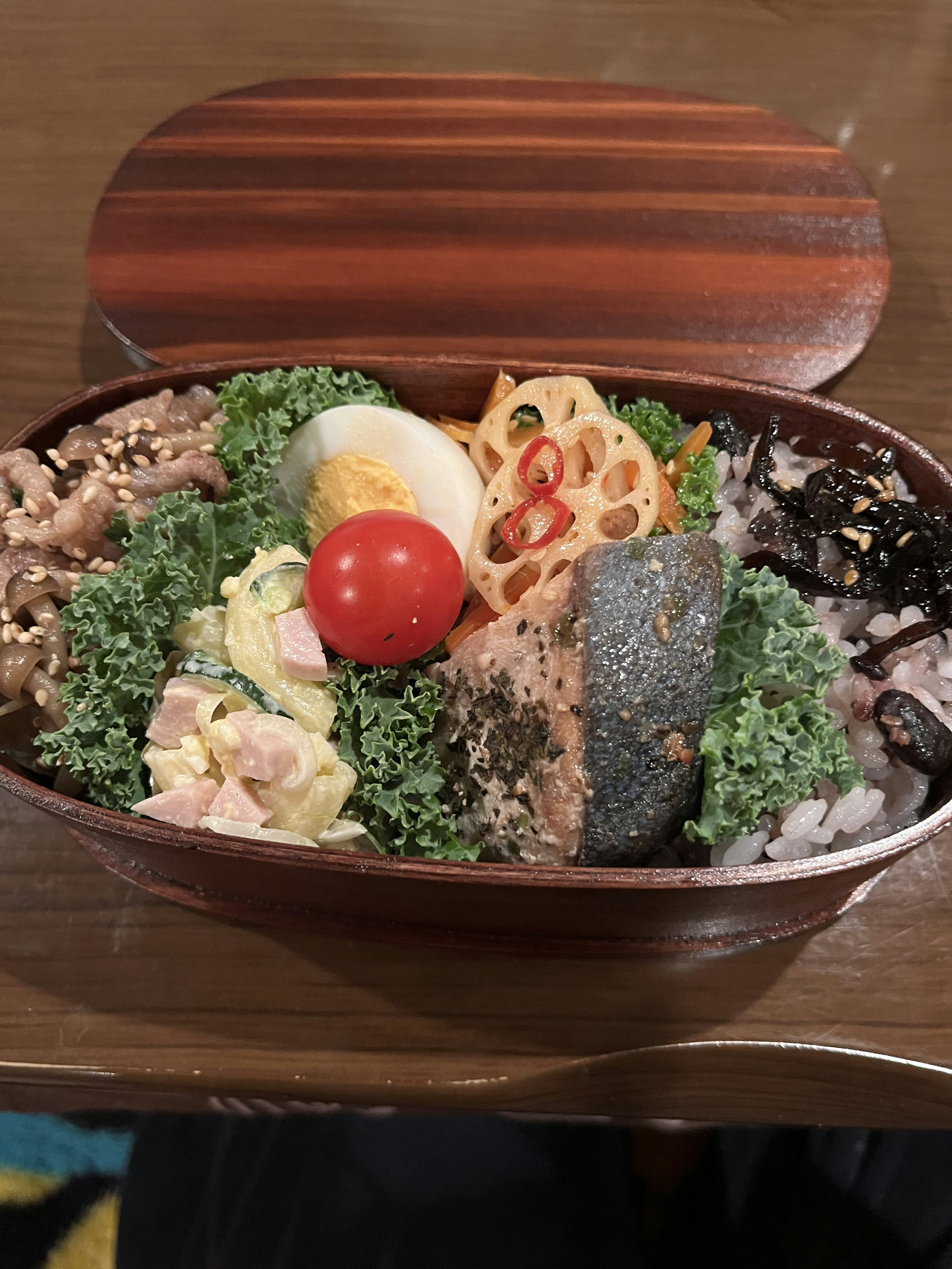 Colorful assortment of dishes in a wooden bento box featuring salmon, boiled egg, lotus root, and salad