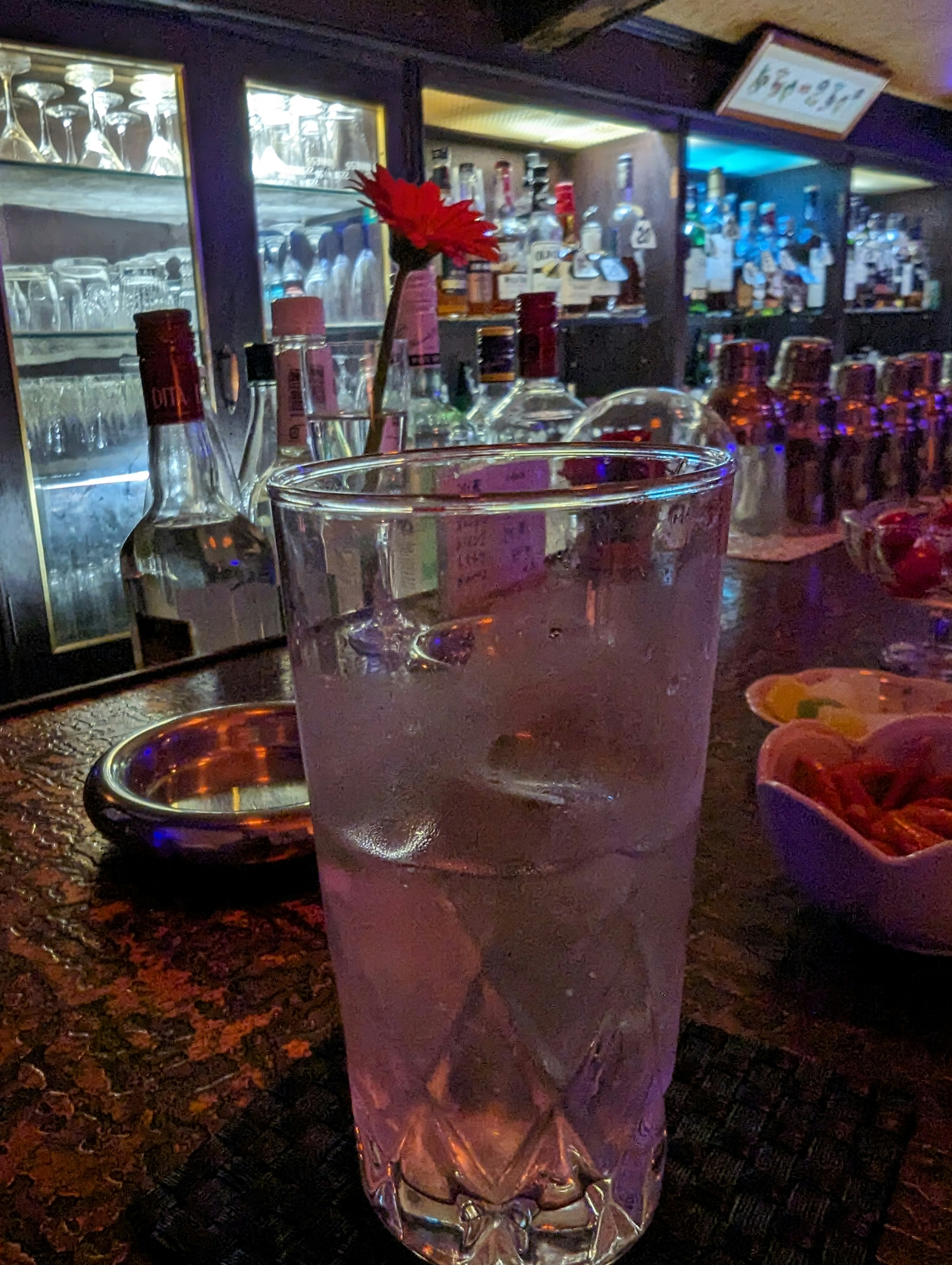 Cocktail glass with a red flower floating and a clear drink Bar background with bottles and lighting