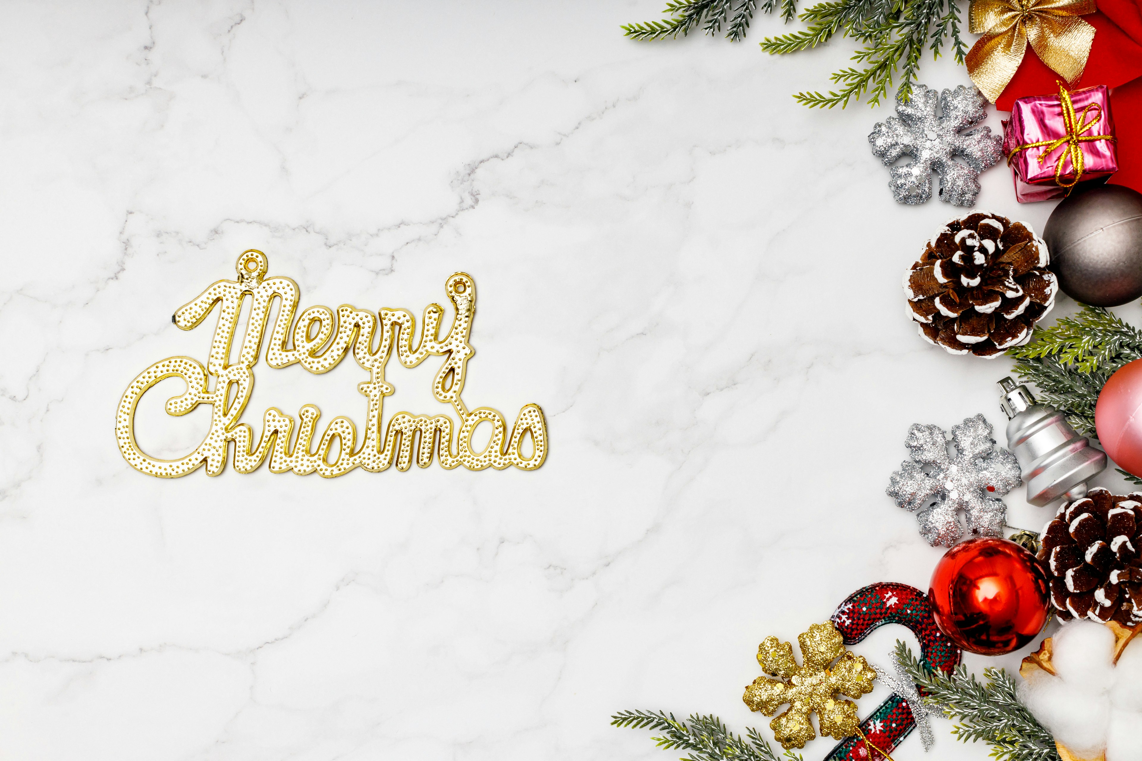 Gold Merry Christmas text on a white marble background with red ornaments and pine cones