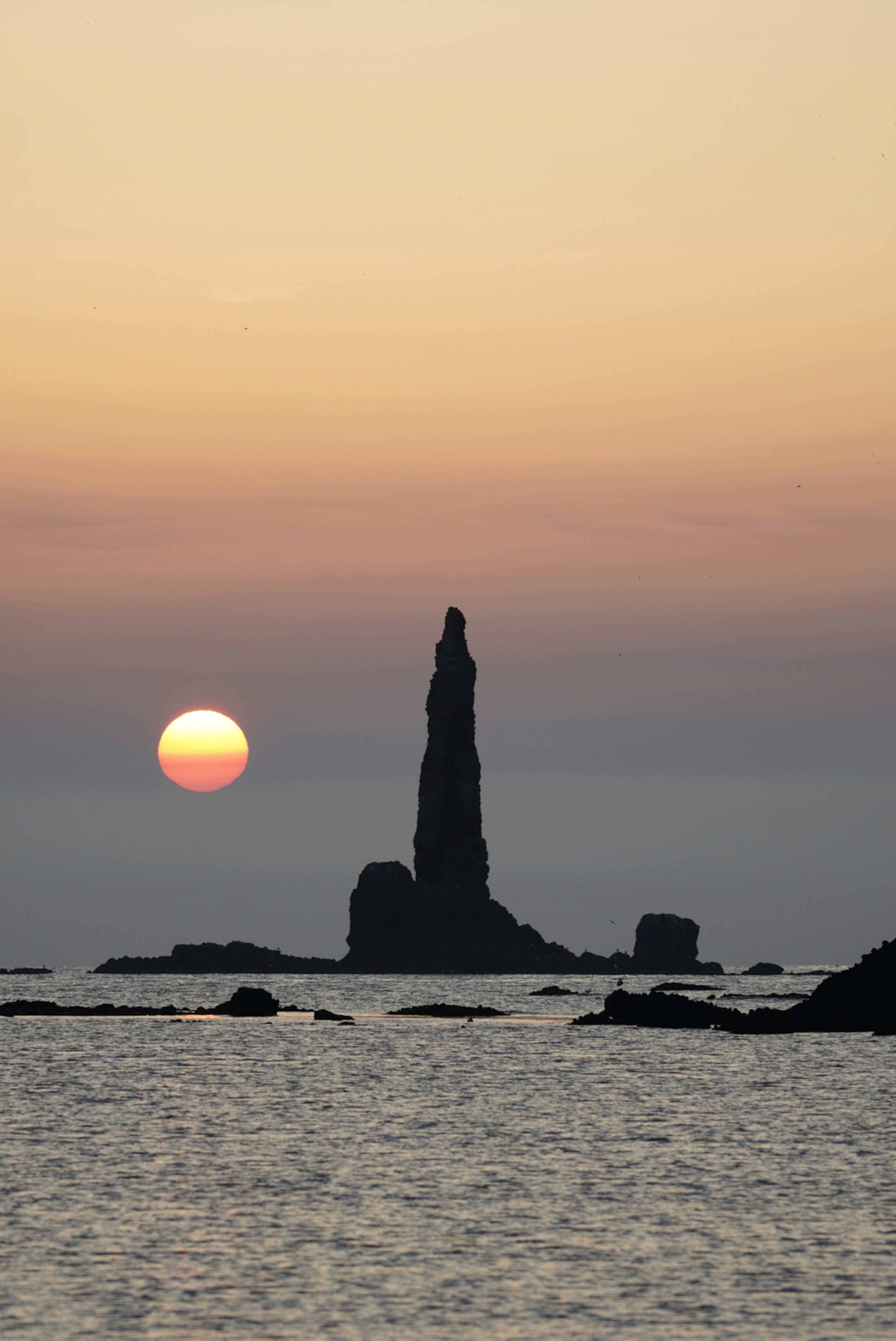 Formación rocosa aislada que se eleva del mar con un atardecer de fondo