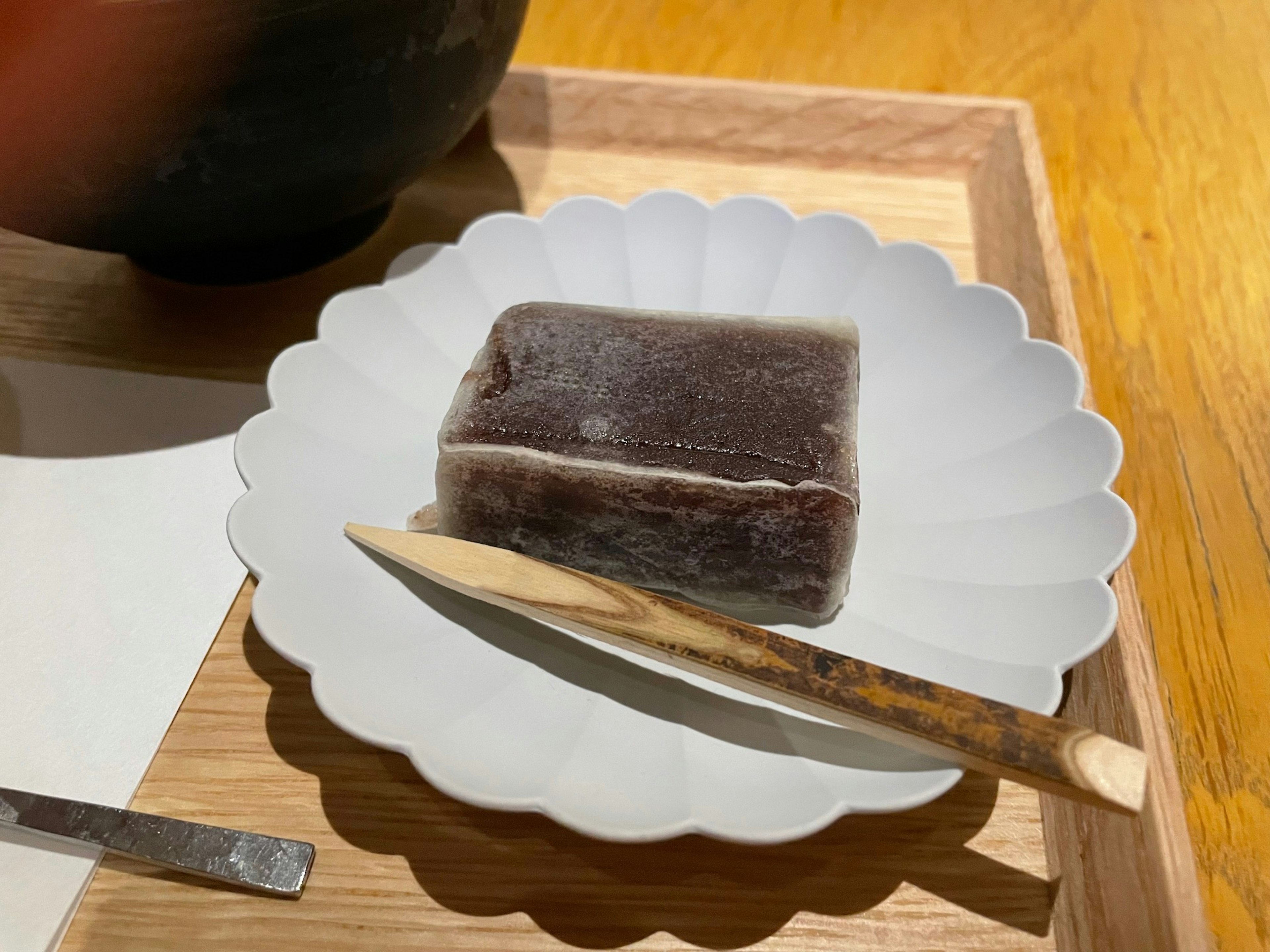 Un morceau de confiserie japonaise sombre sur une assiette blanche accompagné d'une cuillère en bois