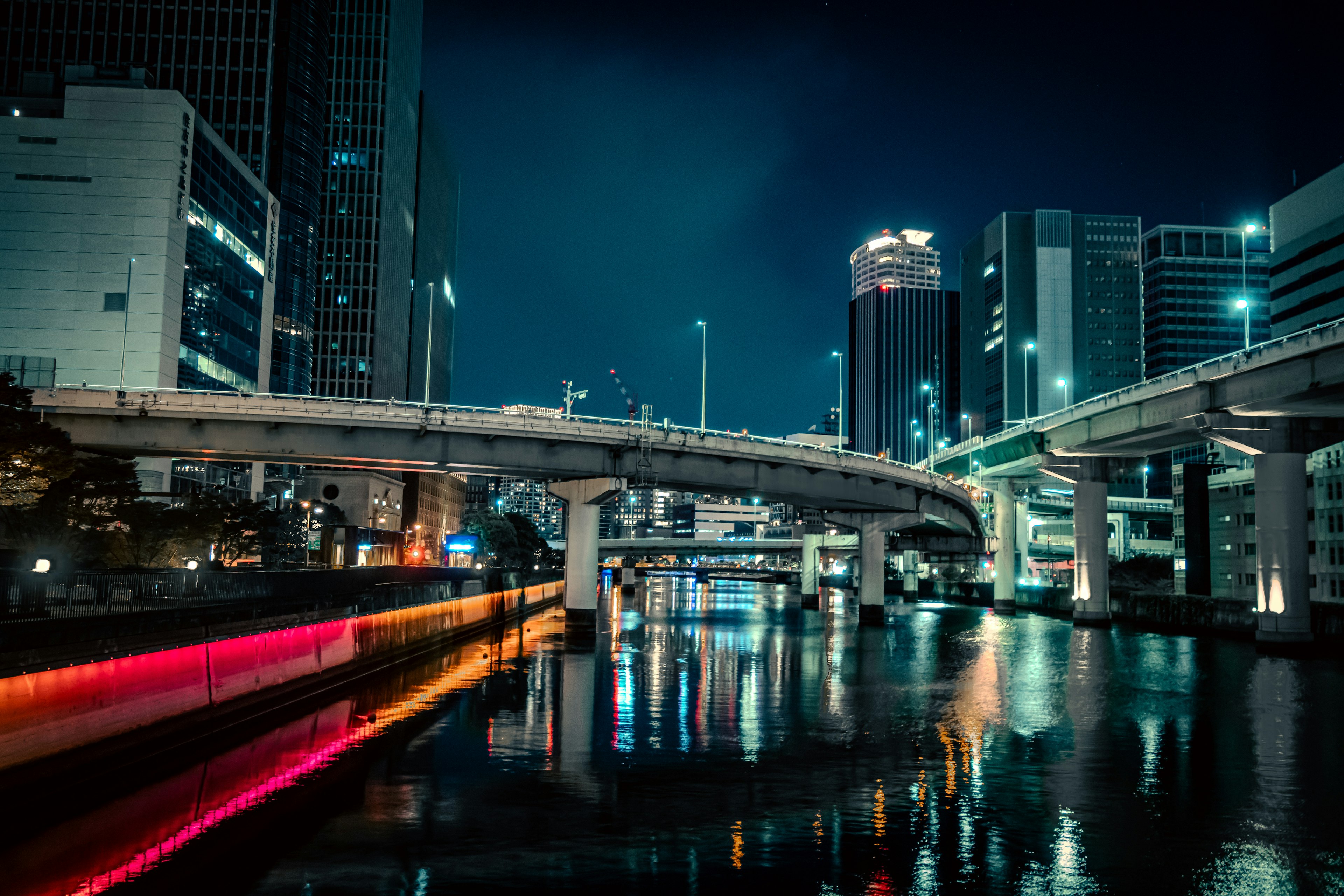 Paisaje urbano nocturno con edificios iluminados reflejados en el río y una autopista arqueada