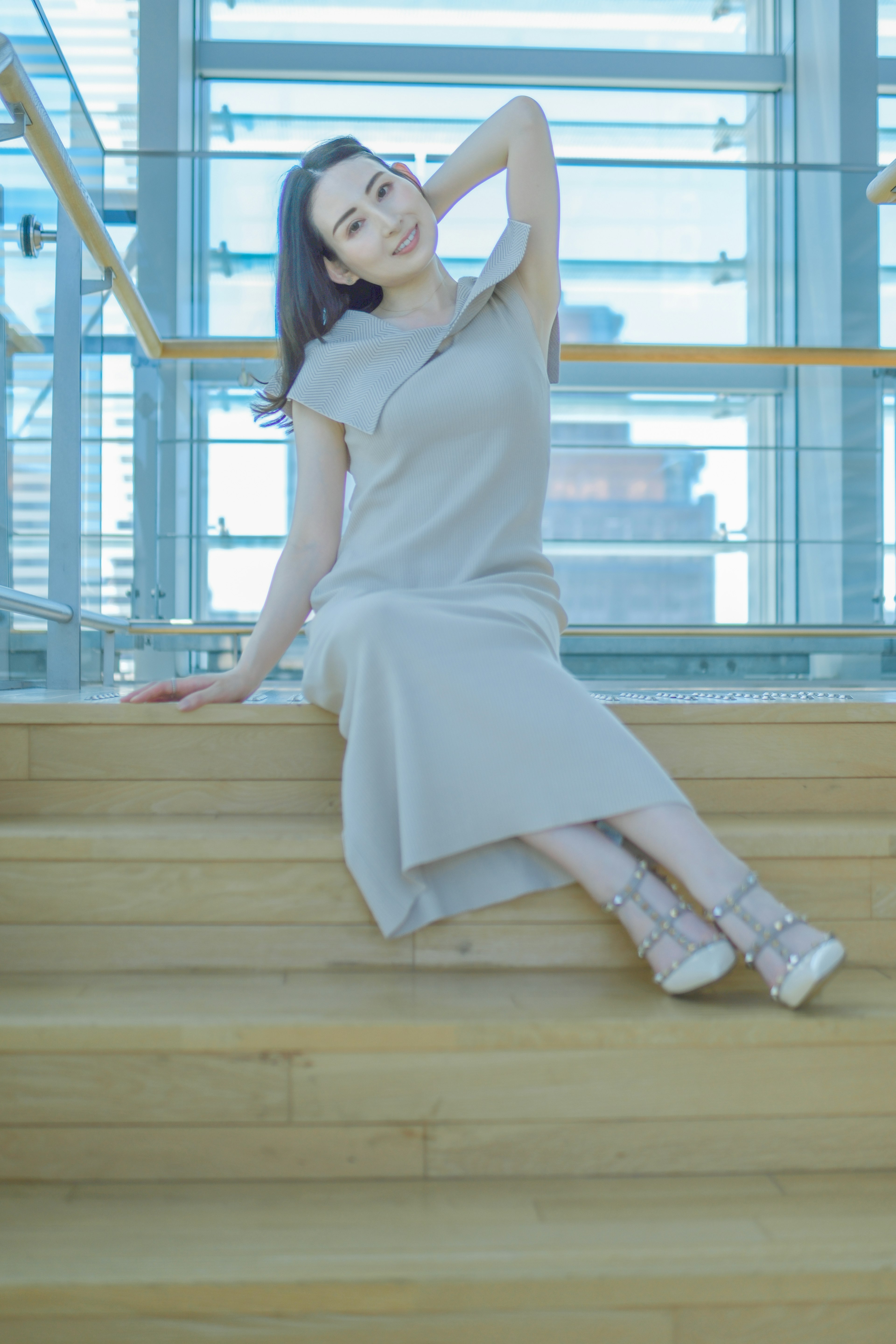 A woman sitting in a bright space wearing a gray dress on wooden stairs in a relaxed pose