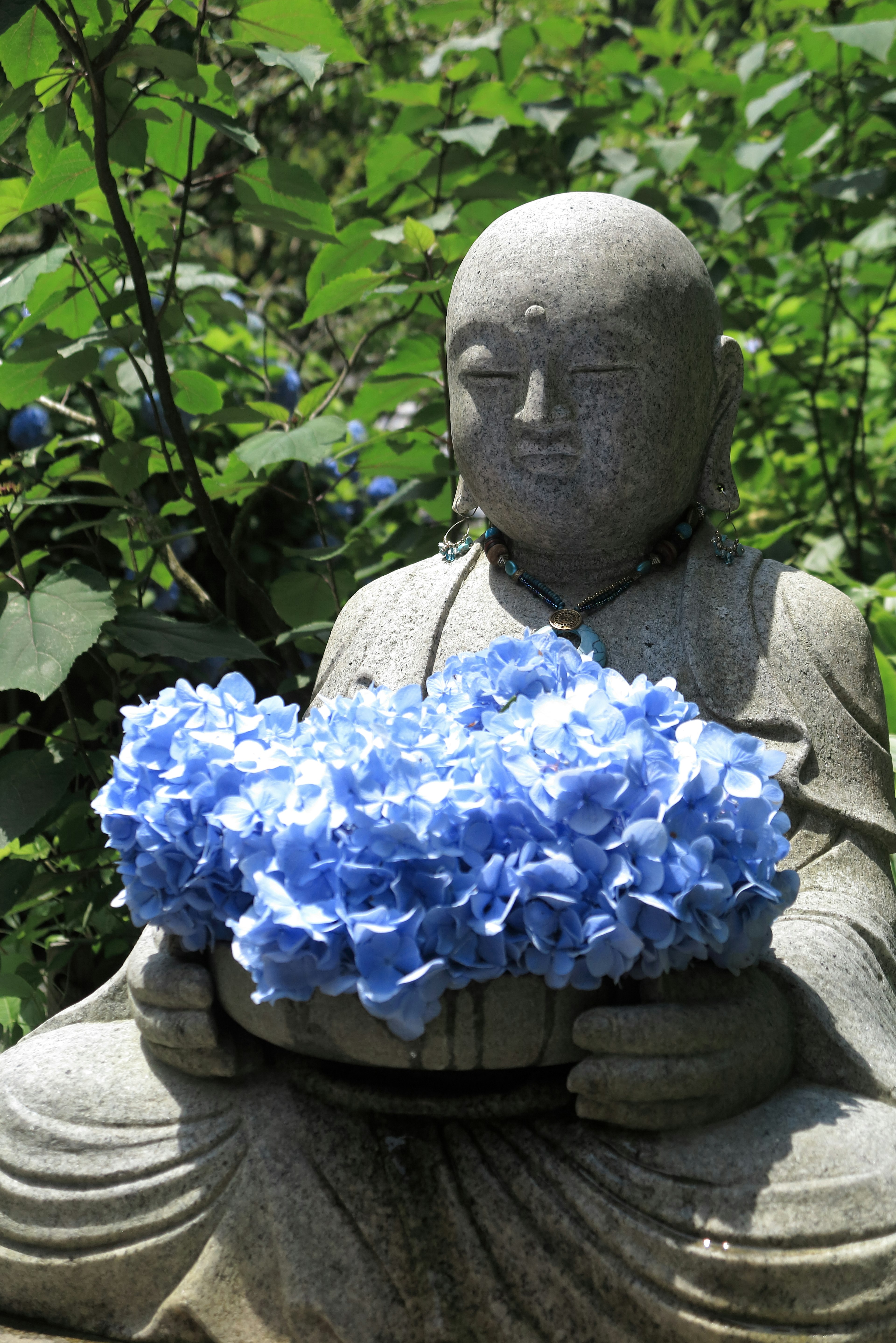 Estatua de Buda de piedra sosteniendo un ramo de flores azules rodeada de vegetación