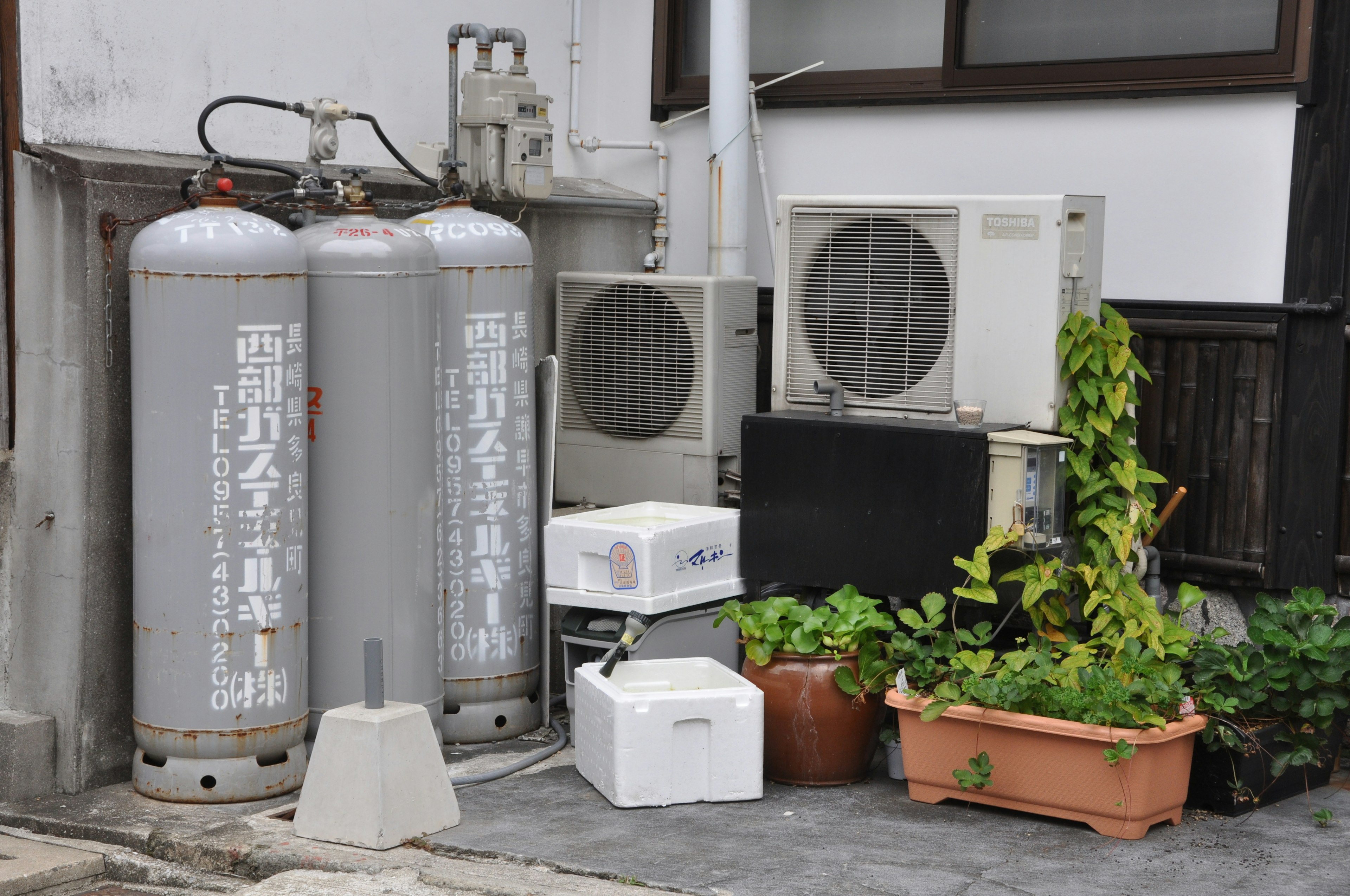 Gray gas cylinders and air conditioning units positioned against a wall in an urban setting