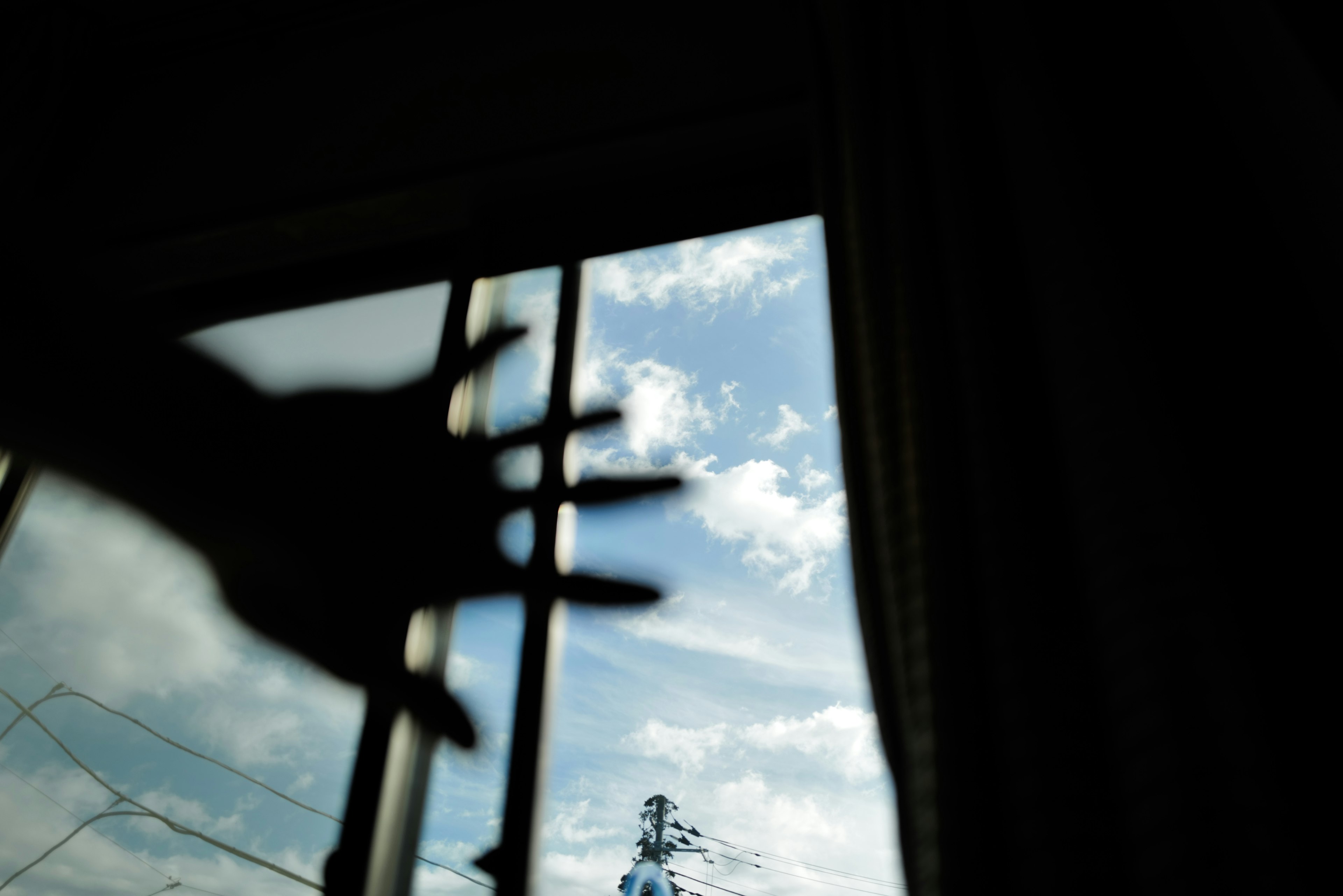 Eine Hand, die nach Fensterstäben greift, mit blauem Himmel und Wolken draußen sichtbar