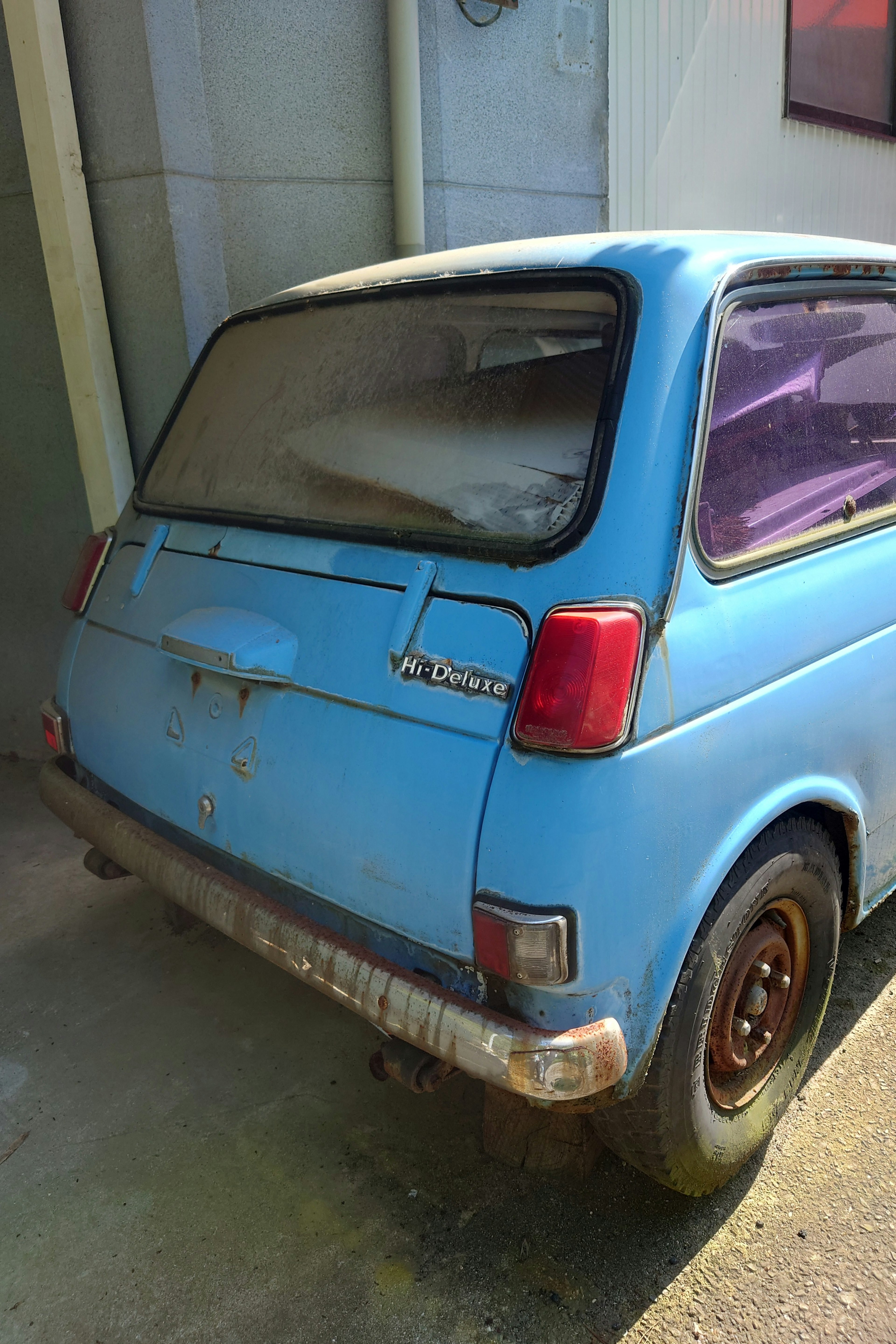 Rear view of an old blue compact car with a dusty window and rusted body