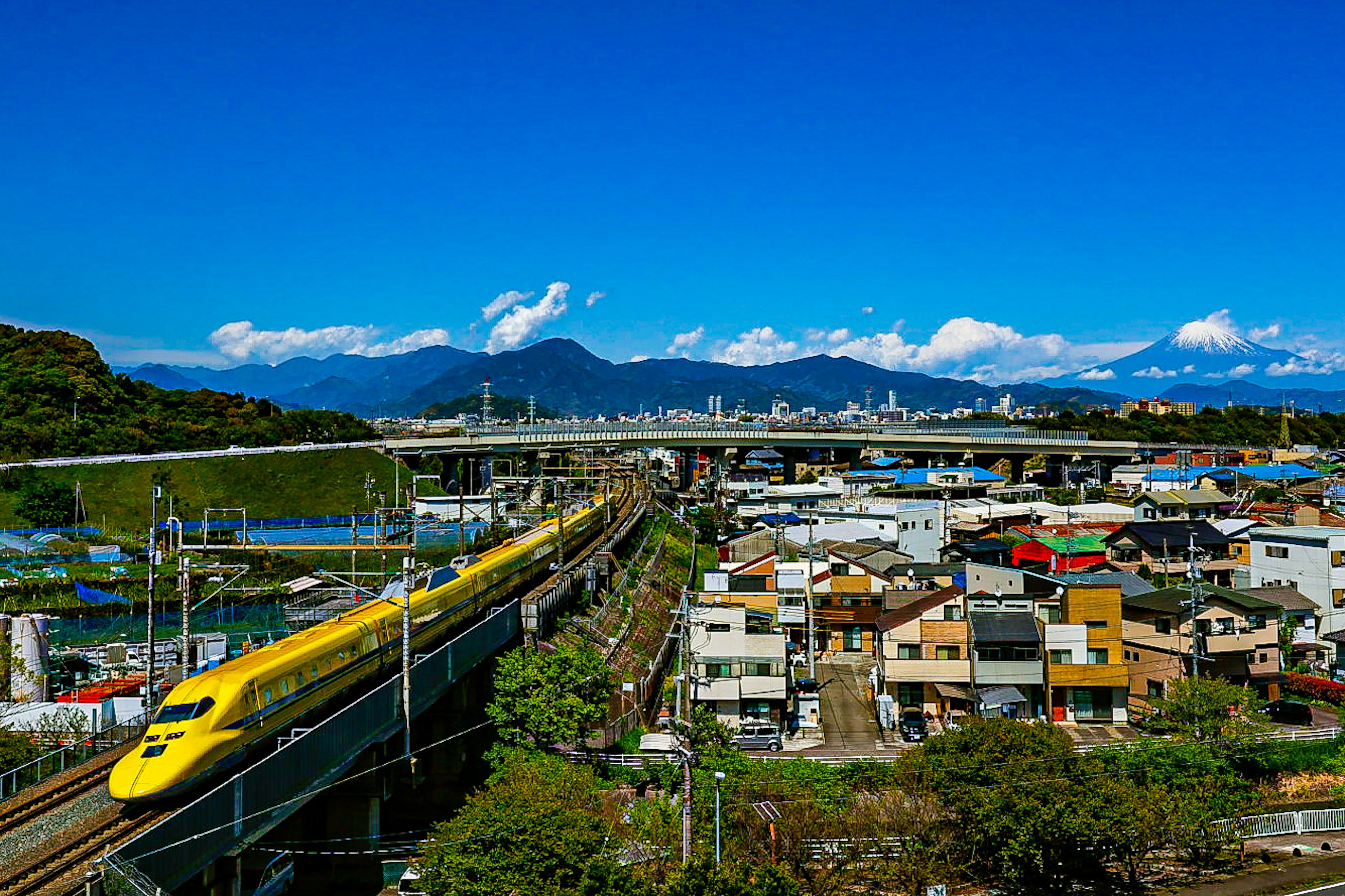 Treno Shinkansen che viaggia sotto un cielo blu con montagne sullo sfondo e paesaggio urbano