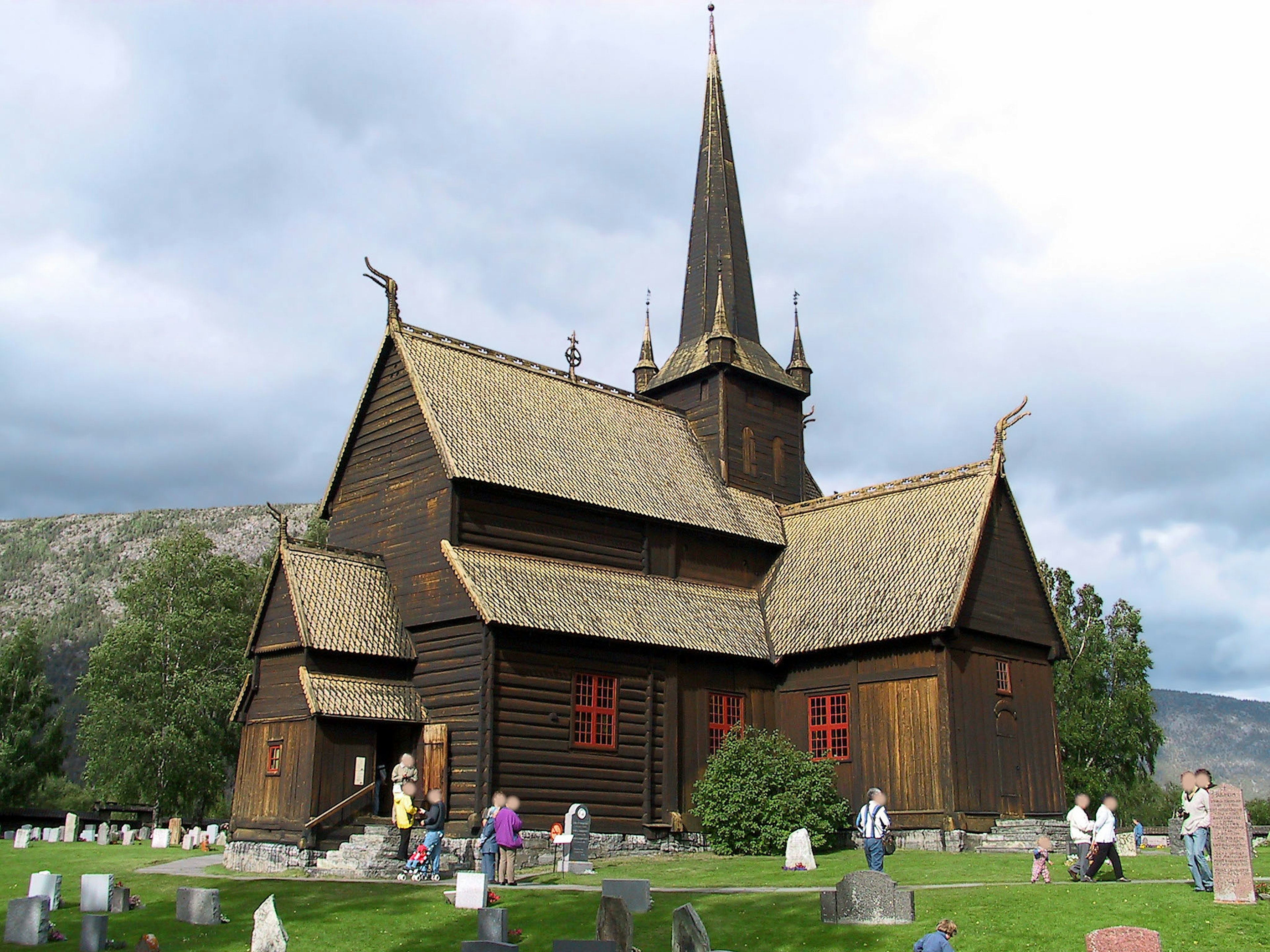 Wooden church with red windows and a spire surrounded by a graveyard and people