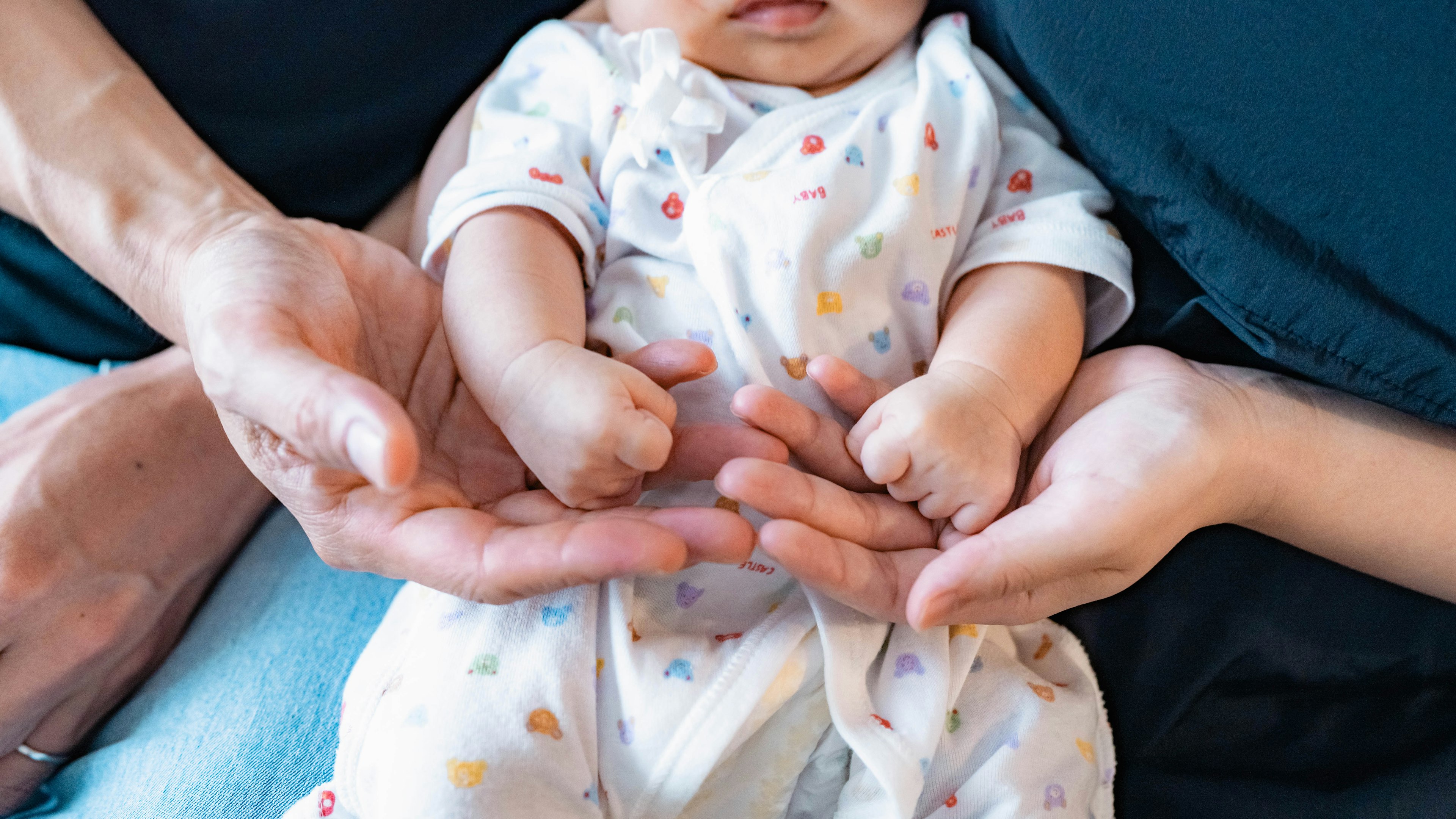 Ein Nahaufnahmebild eines Babys, umgeben von Erwachsenenhänden Das Baby trägt einen bunten Overall