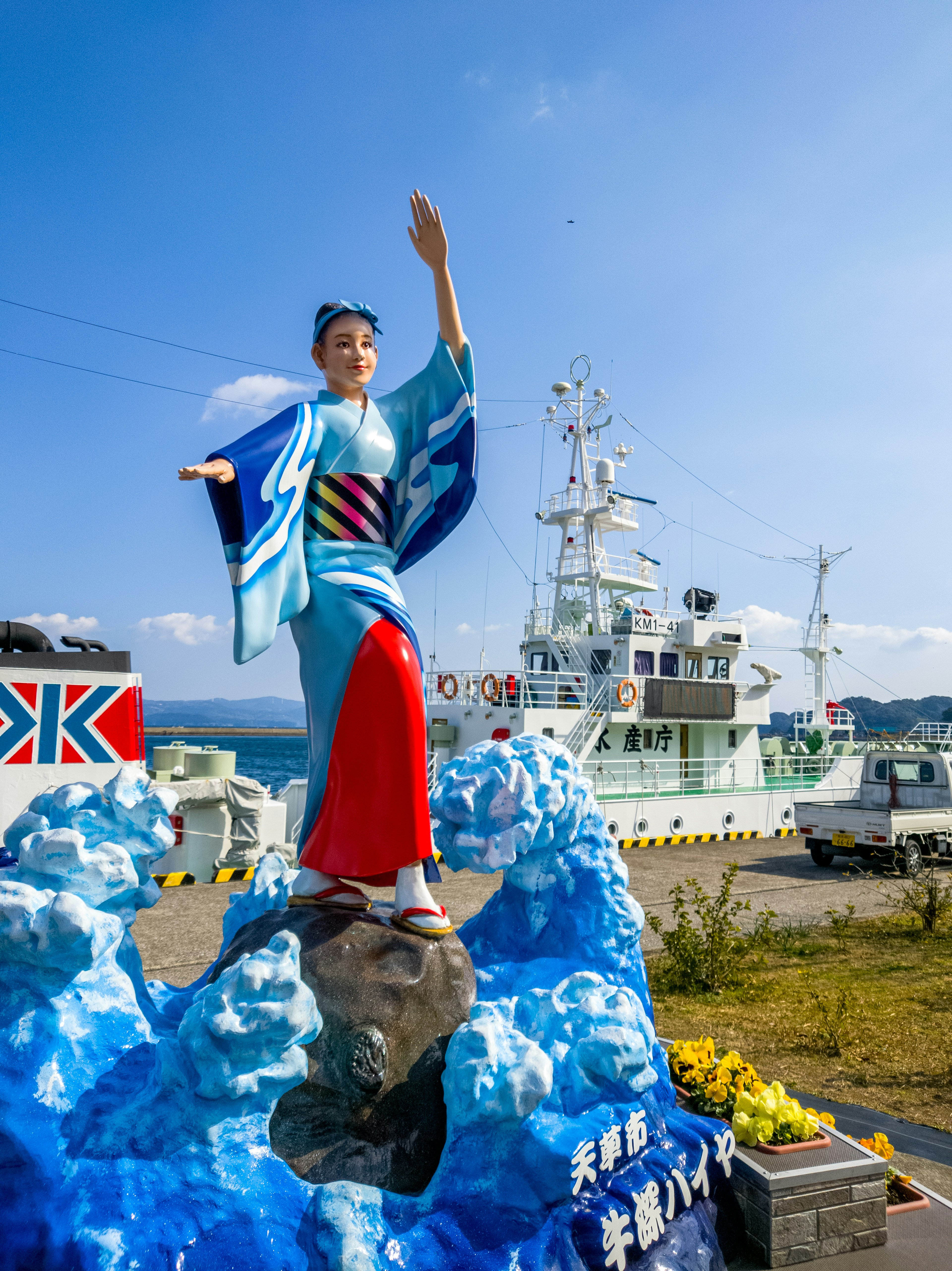 Statue einer Frau in Kimono, die auf blauen Wellen steht, mit einem Schiff im Hintergrund